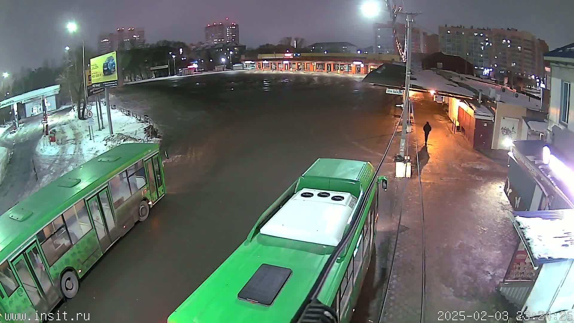 Two green buses are parked in a wet, snow-covered bus station at night, with a person walking in the background.