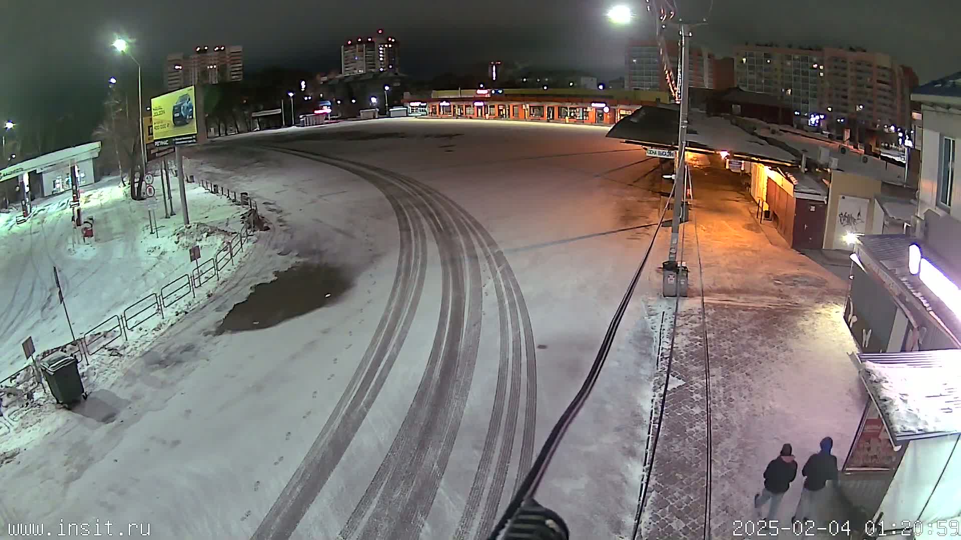 A snow-covered nighttime parking lot with tire tracks, buildings in the background, and two people walking.