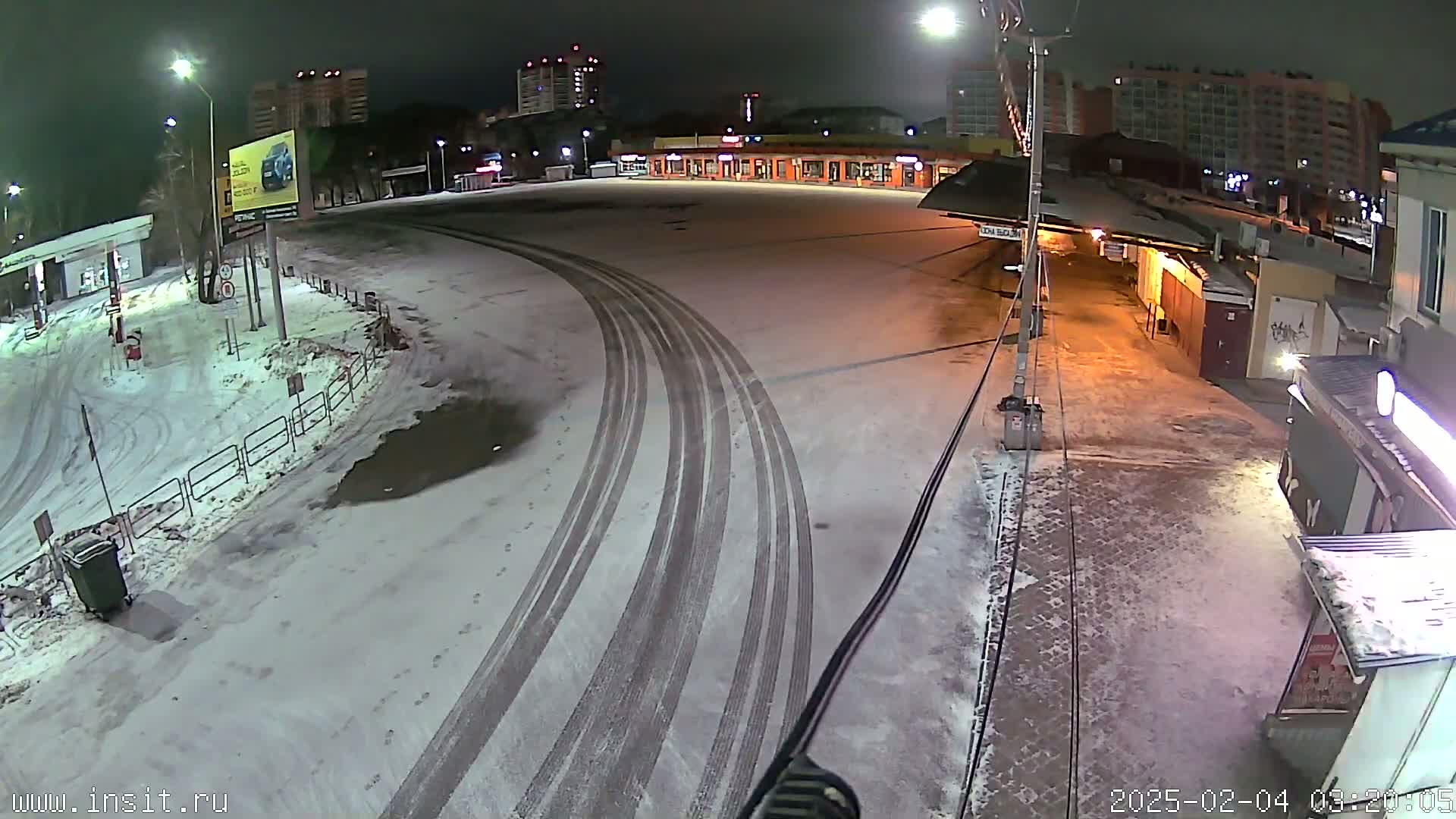A snow-covered nighttime parking lot shows tire tracks curving through the area near buildings, with some lights illuminating the scene.