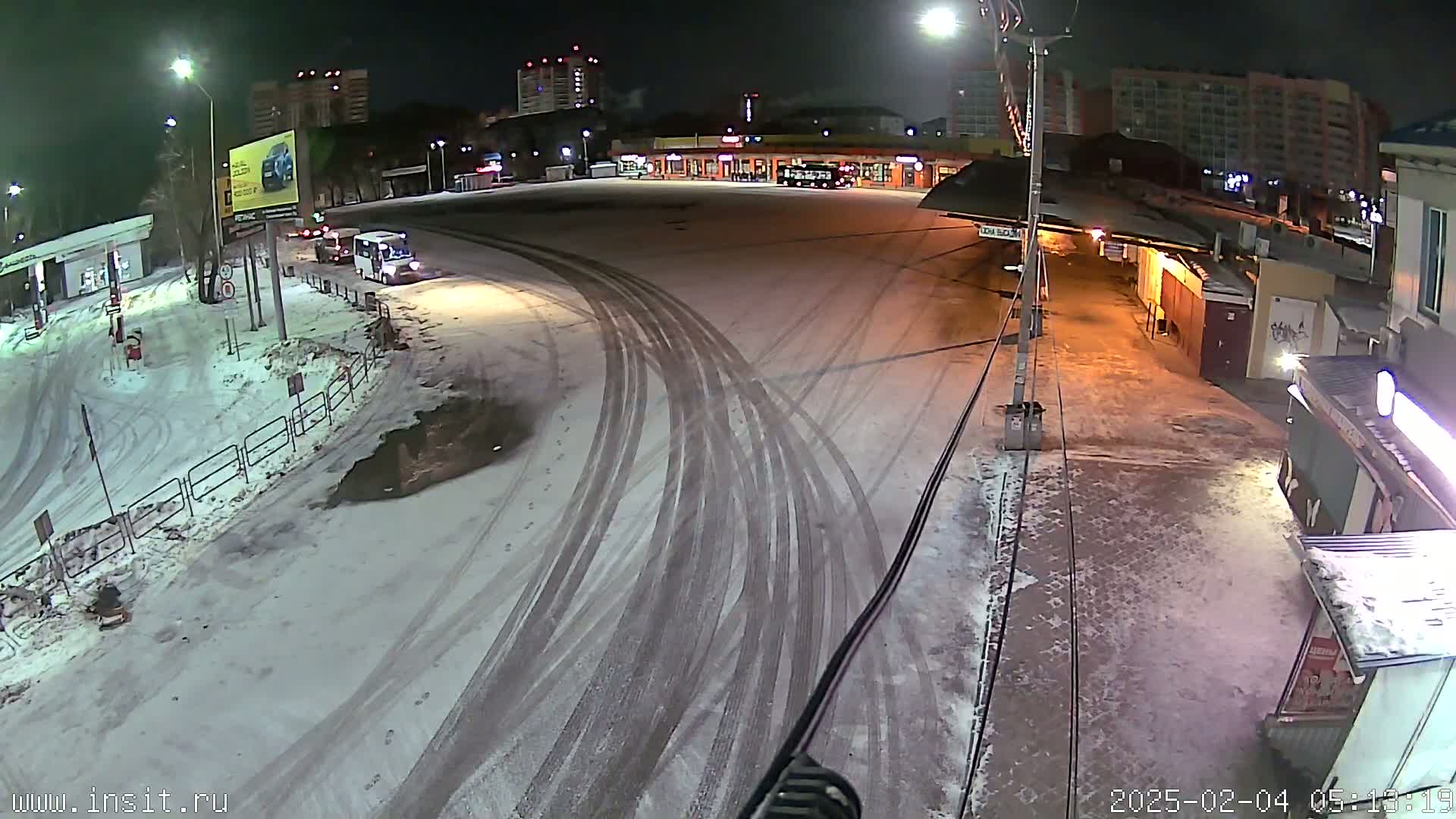 A snow-covered nighttime scene shows a bus station and surrounding area with tire tracks in the snow.