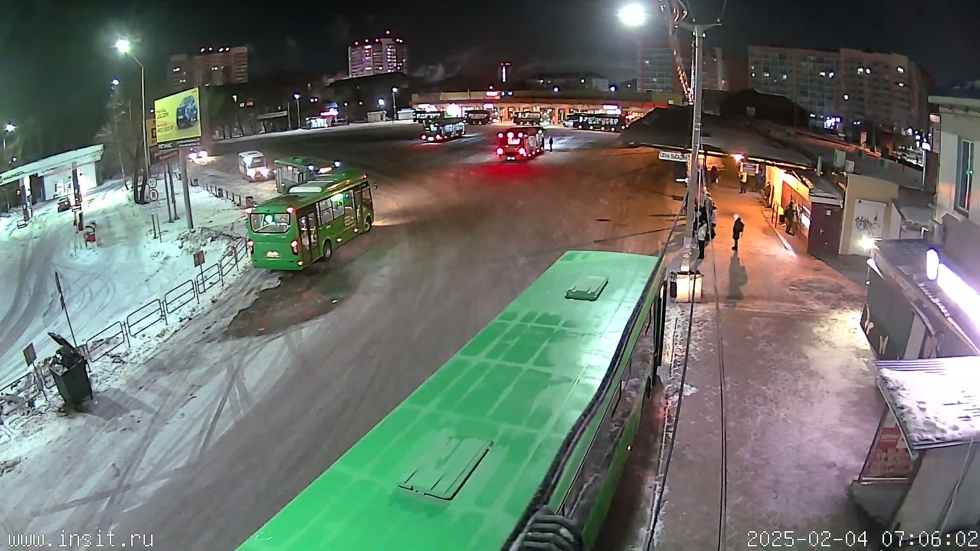 A nighttime view of a snowy bus station with several buses, some people walking, and buildings in the background.