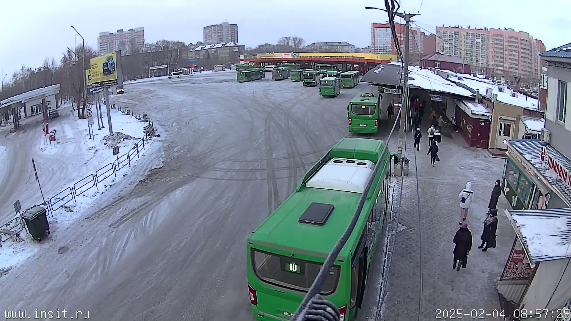 A snowy bus depot with numerous green buses and several pedestrians near a small building.