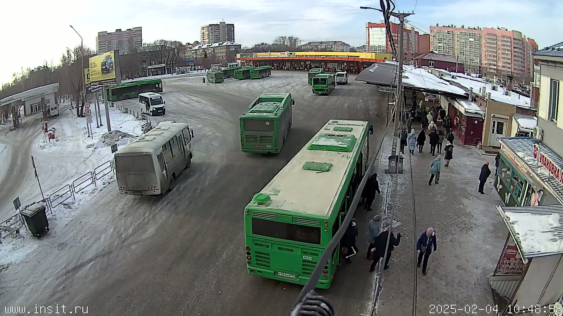 A bus station with several buses, some people waiting, and snow on the ground under a partly cloudy sky.