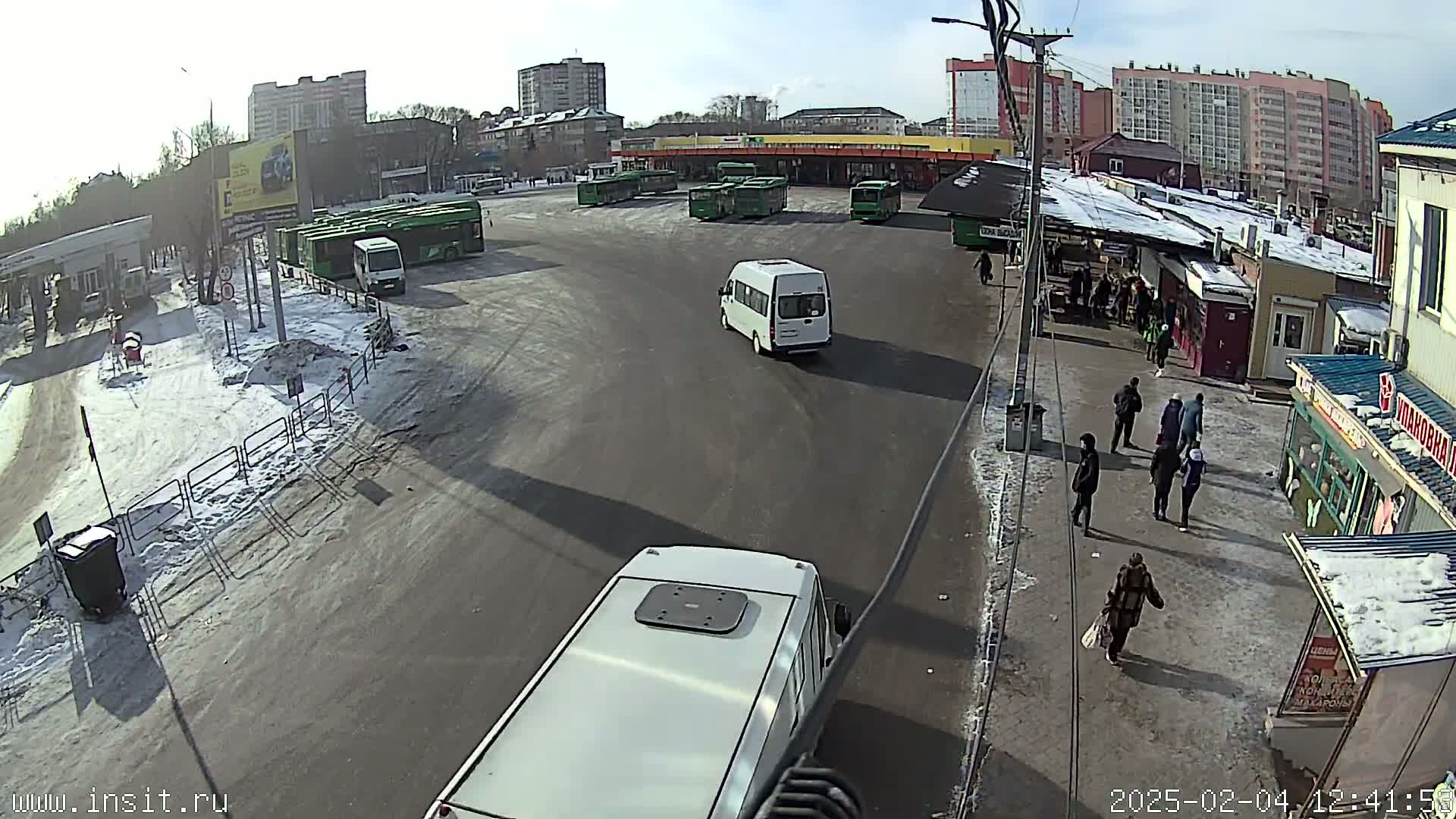 A bus station with several buses and vans, some pedestrians, and patches of snow on the ground under a sunny sky.