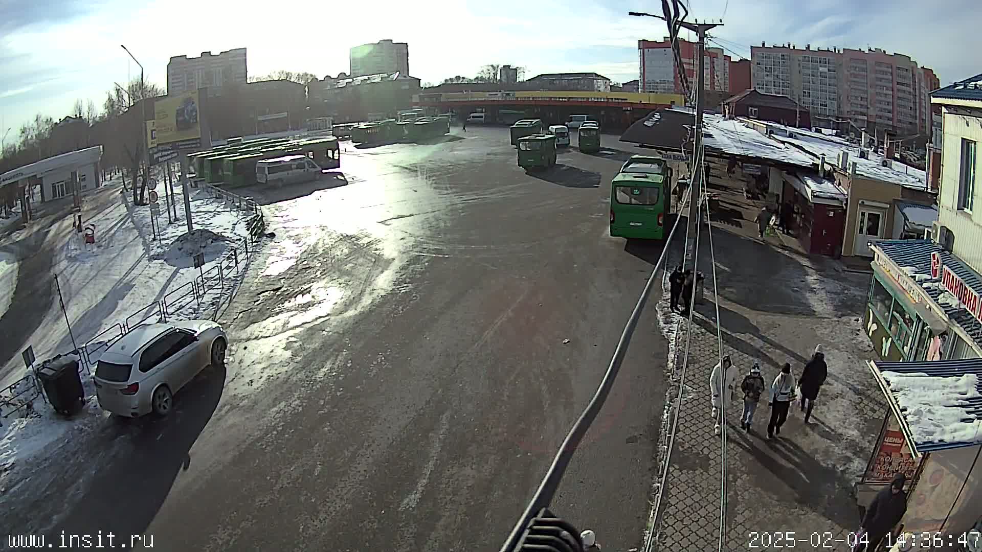A sunny day at a bus station with melting snow and ice on the ground, multiple buses parked, and several people walking around.