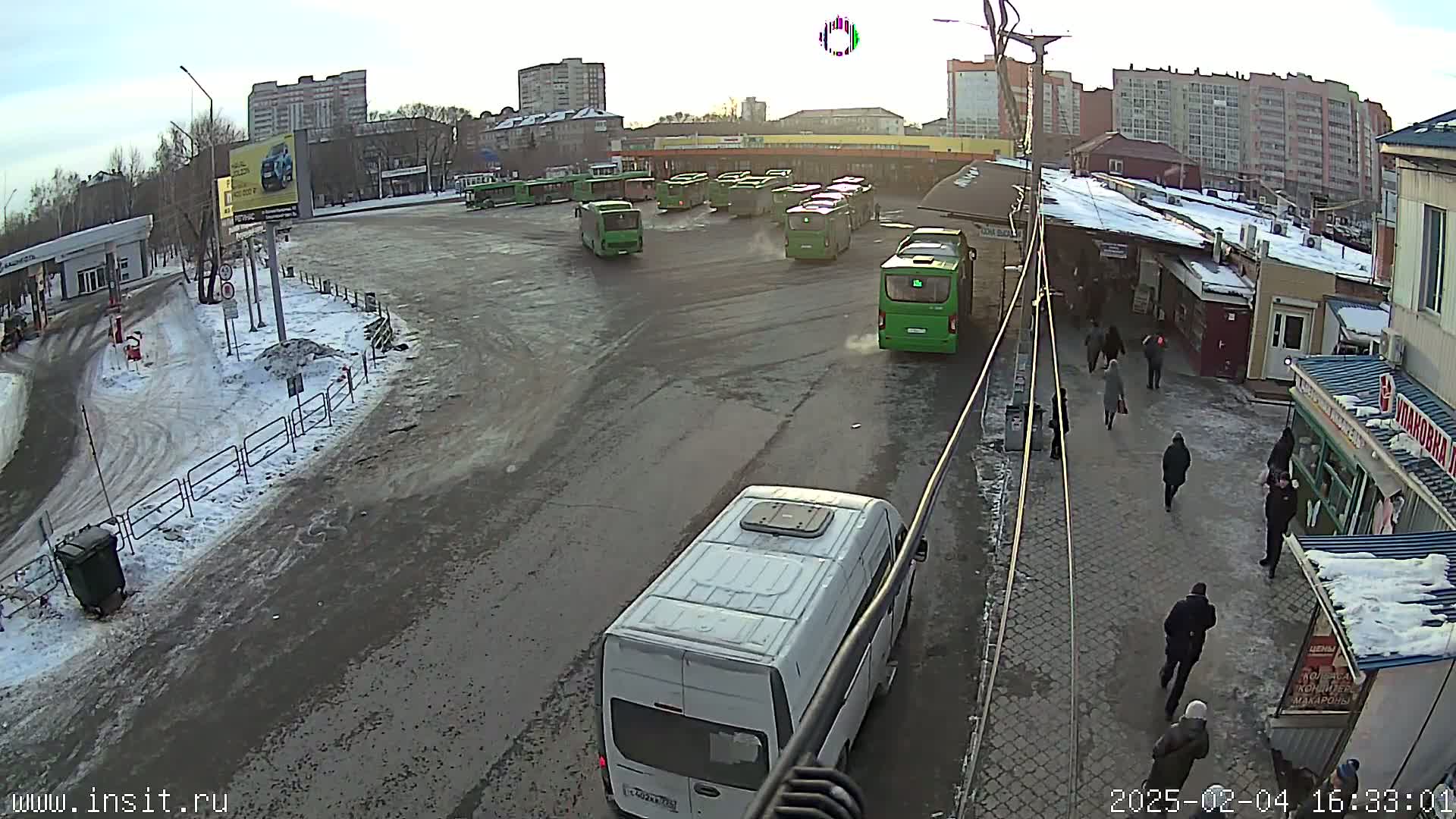 A bus station with numerous green buses and a white van, surrounded by buildings and people walking on a snow-covered, paved area in overcast conditions.