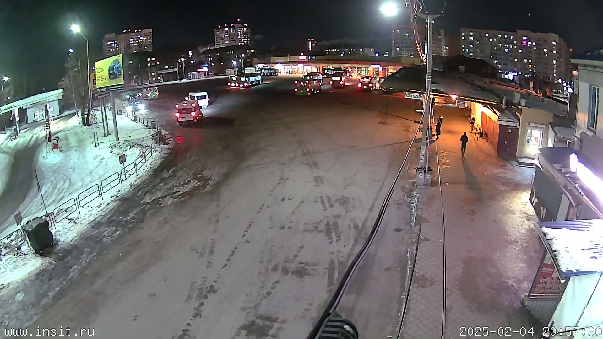 A snow-covered nighttime street scene shows several vehicles near a bus stop, with pedestrians walking nearby, in front of multi-story apartment buildings.