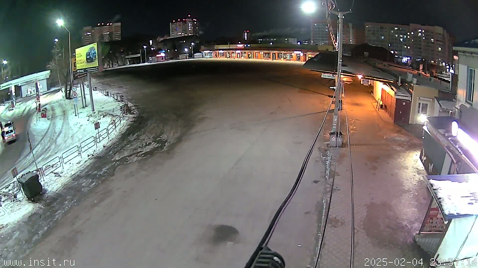 A mostly empty, snow-dusted parking lot at night, bordered by buildings and a street with a few cars, under streetlights.