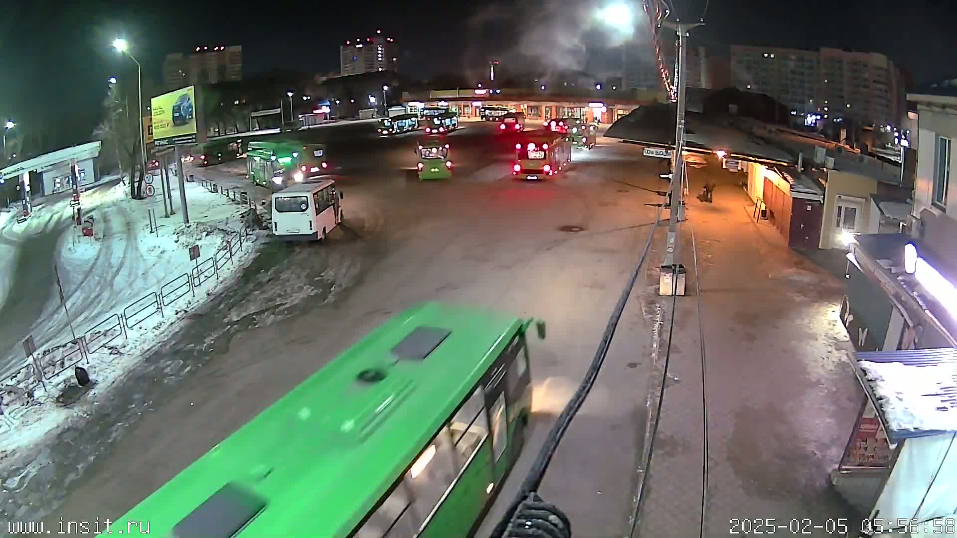 A nighttime view of a bus station with several buses parked and a green bus moving in the foreground, snow on the ground, and buildings in the background.