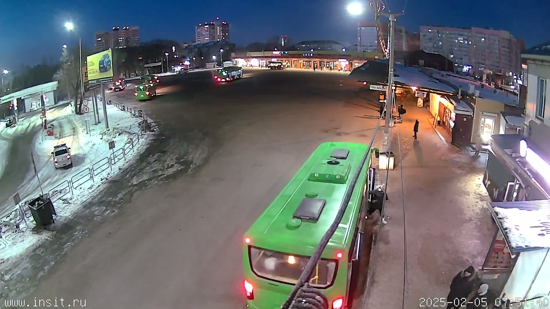 A nighttime scene at a bus station shows a green bus and several other vehicles in a snowy, dimly lit area with several buildings in the background.