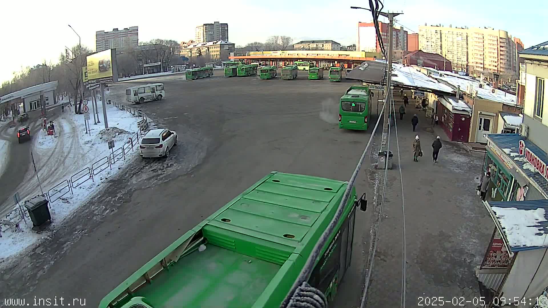 A bus depot with several green buses parked, a few cars, and pedestrians walking around on a partly snowy, overcast day.