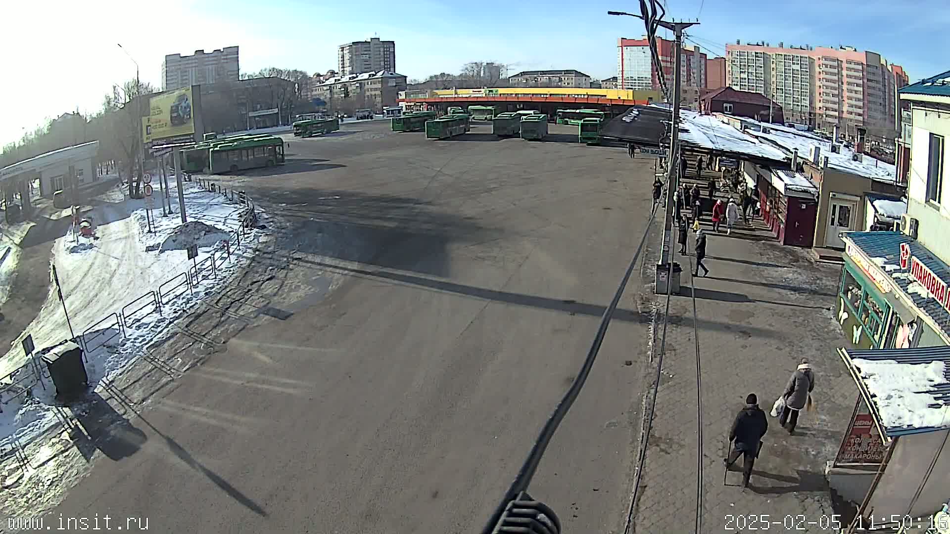 A sunny day at a bus station with several buses parked and a few people walking around, some snow and ice patches visible on the ground.