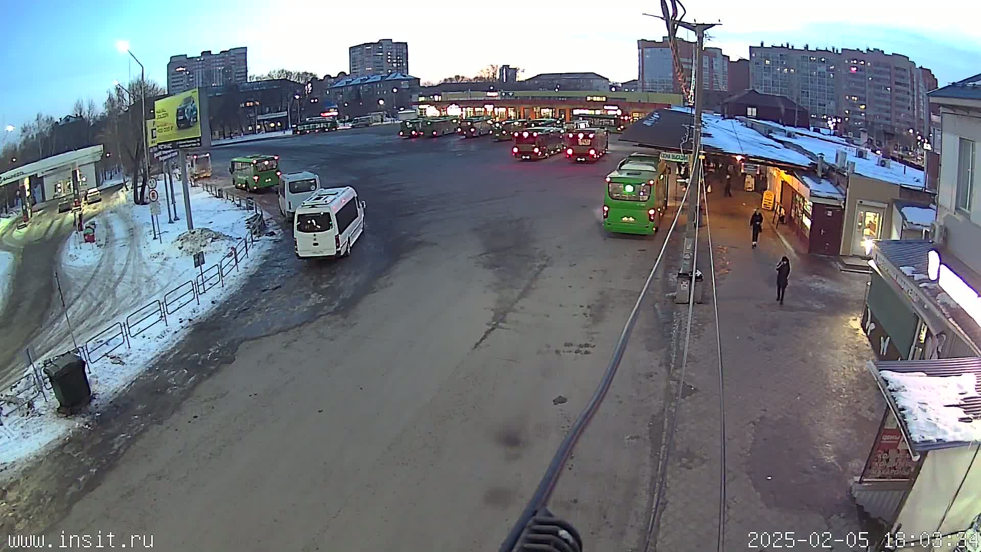 A bus station with numerous buses and a few vans parked in a large lot, partially covered in snow, under a twilight sky.