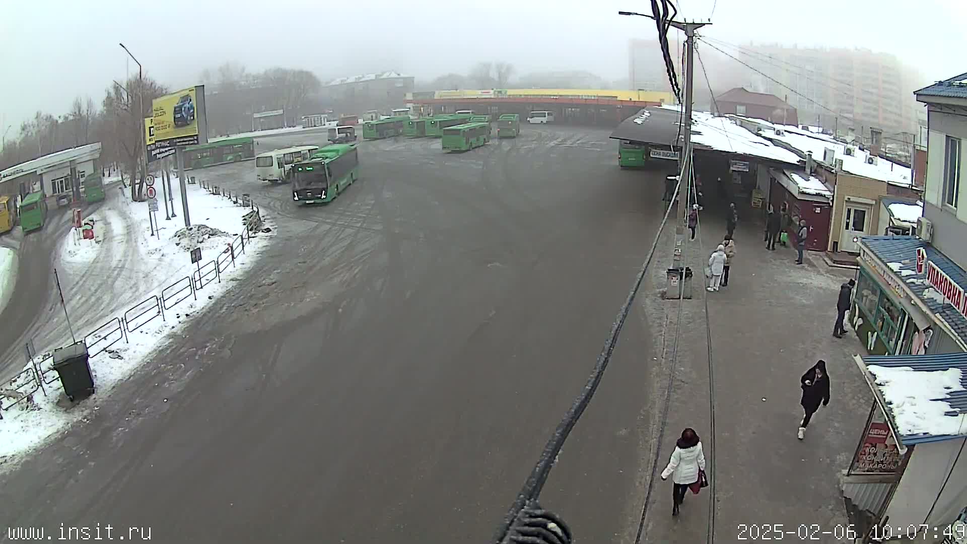 A bus station with numerous buses and a few pedestrians is covered in a light layer of snow in foggy conditions.