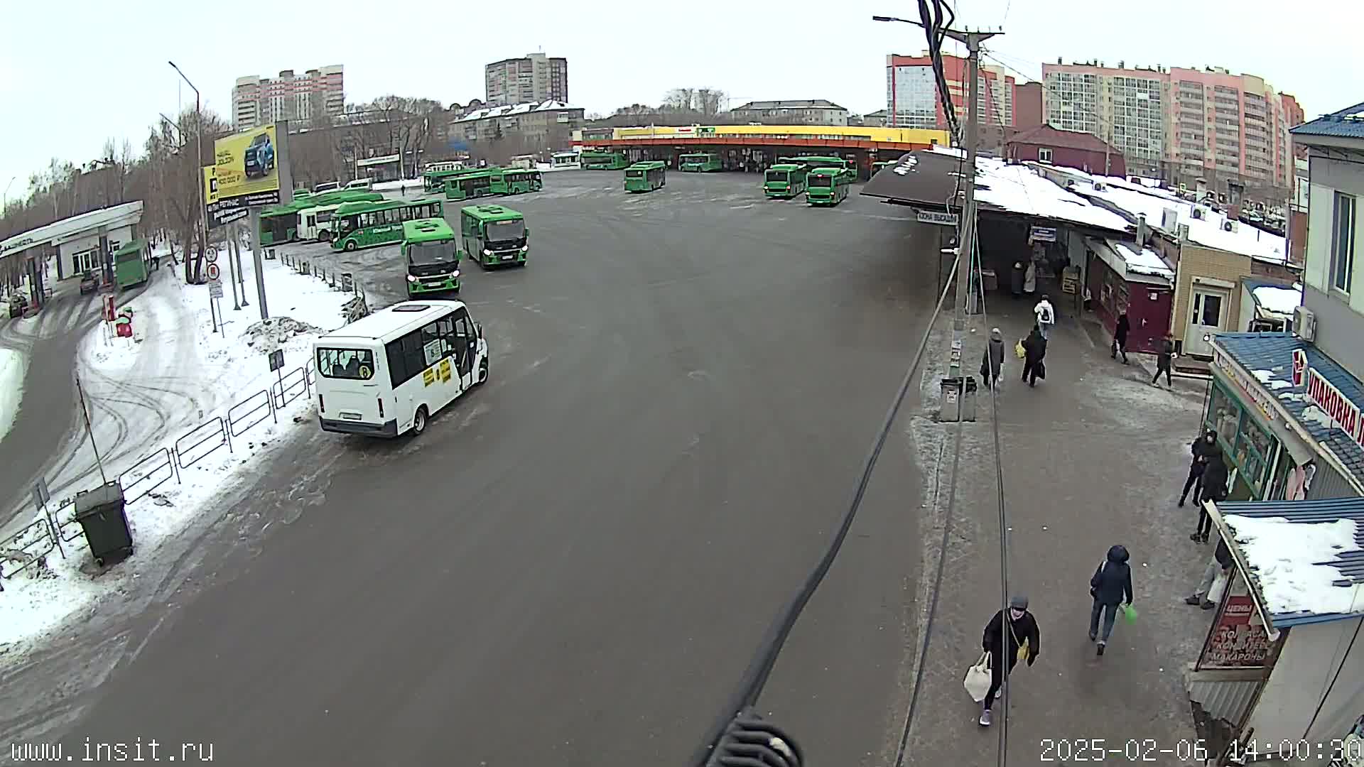 A bus station with numerous green buses and a few pedestrians, partially covered in snow, on an overcast day.