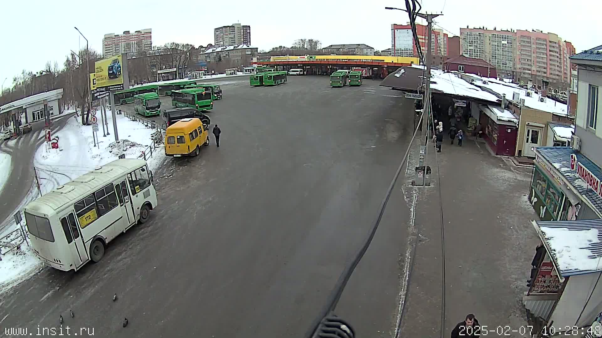 A bus station with several buses parked, some cars, and pedestrians, on a snowy day.