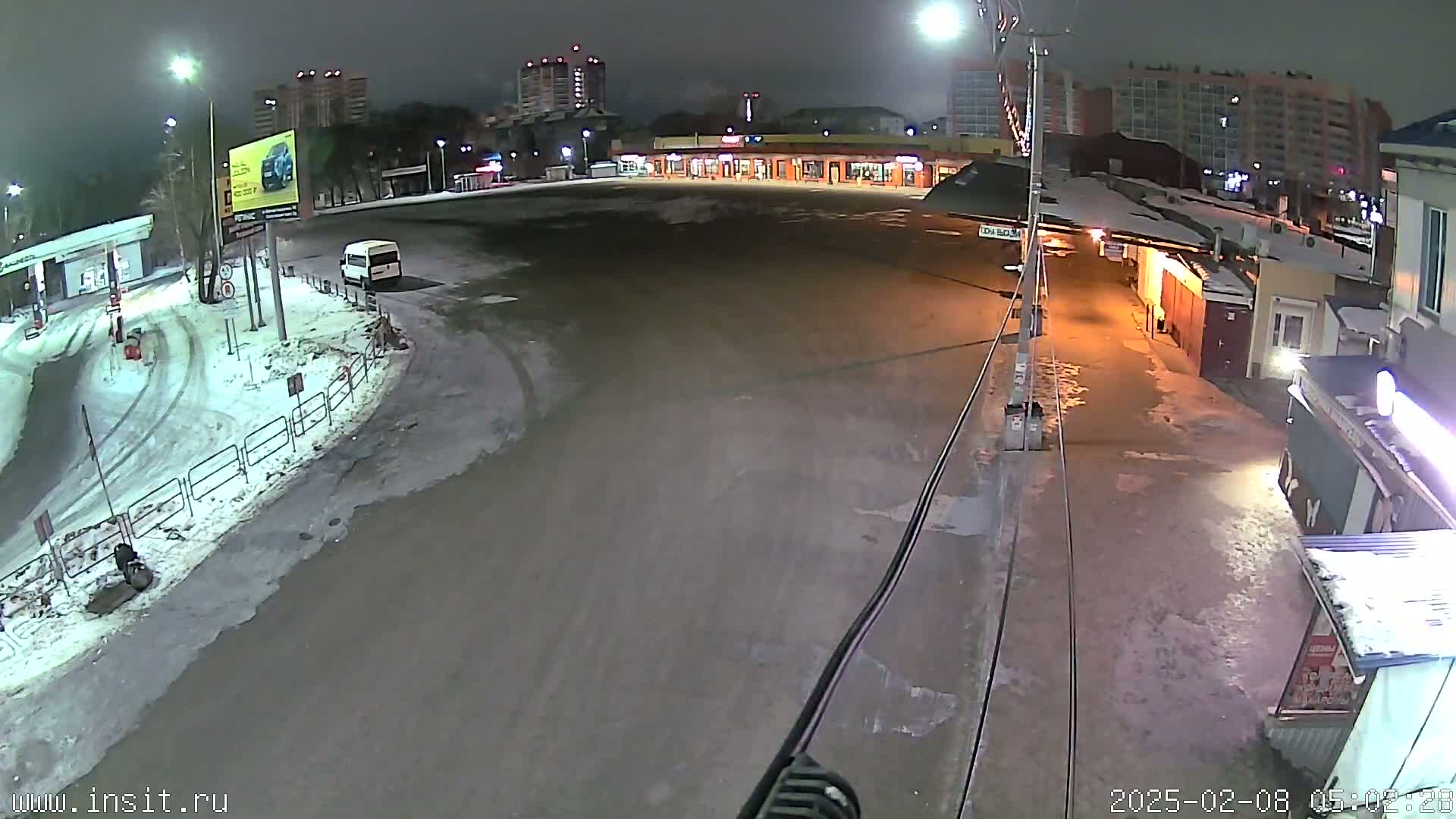 A mostly empty, snow-dusted parking lot at night, with a few buildings and a van visible, under dark skies.