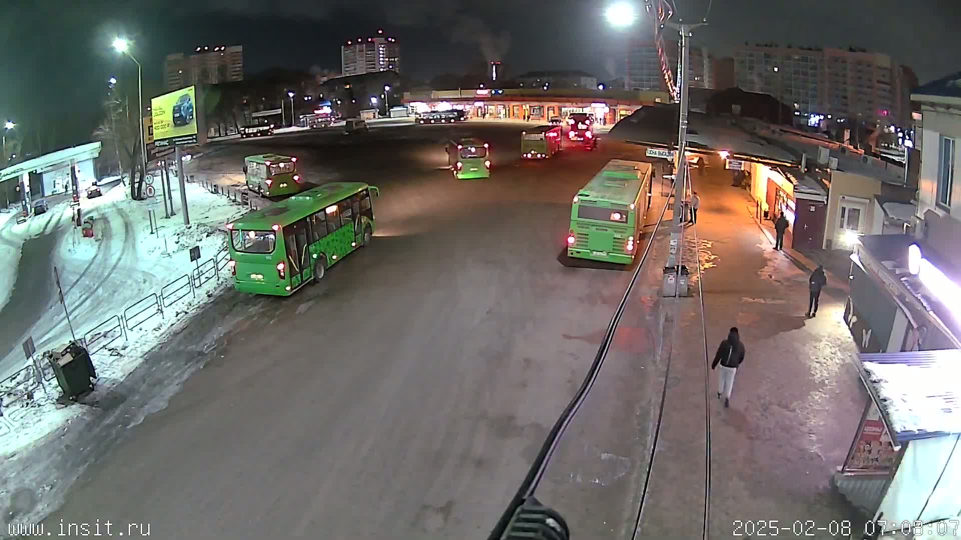 Several green buses are parked in a snowy bus depot at night.