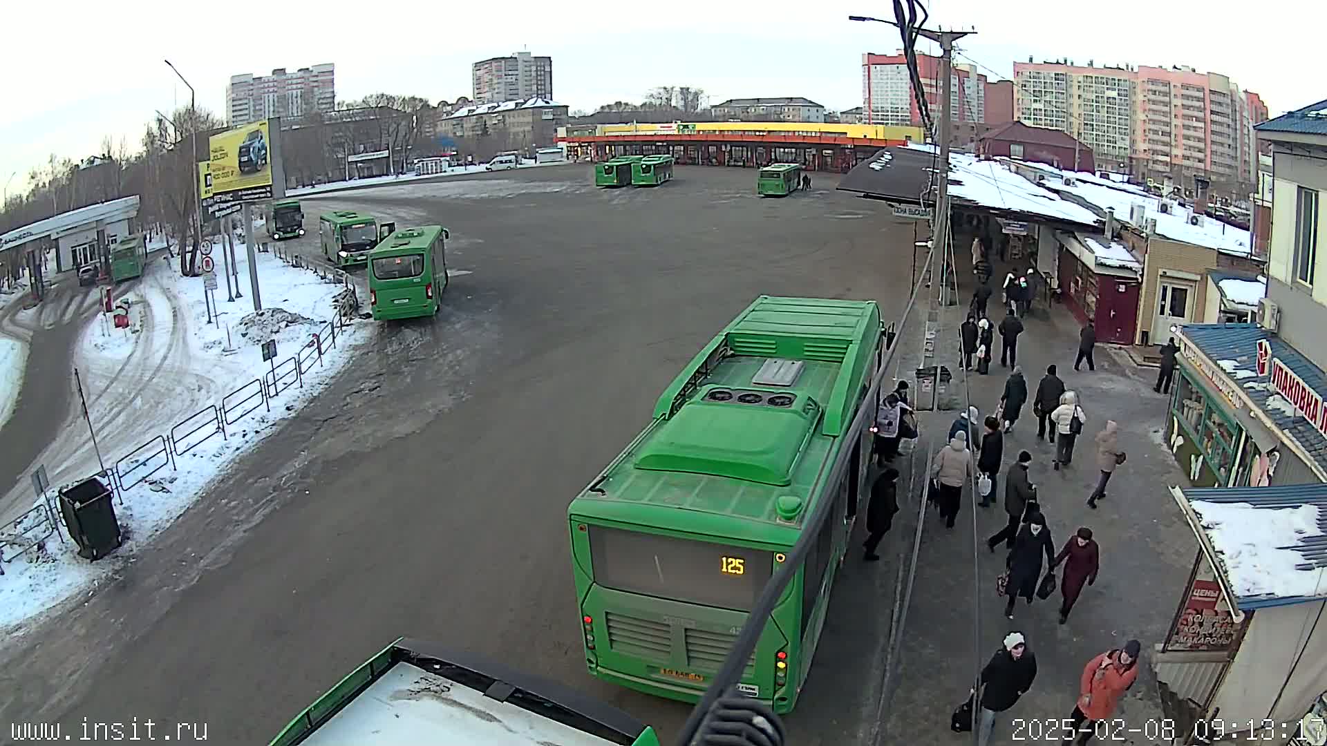 A bus station with several green buses and numerous people milling around on a snowy, overcast day.