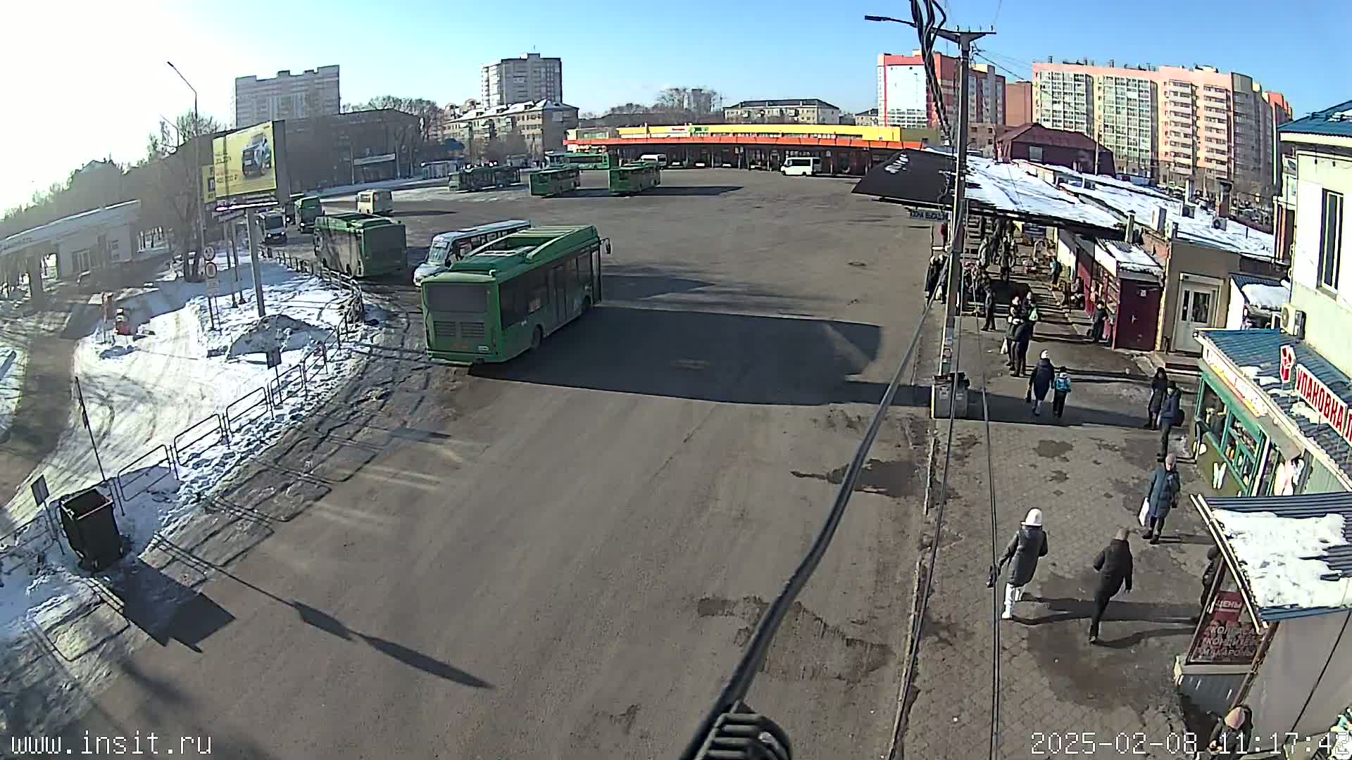 A bus depot with several buses parked, surrounded by buildings and people, on a sunny day with patches of snow on the ground.