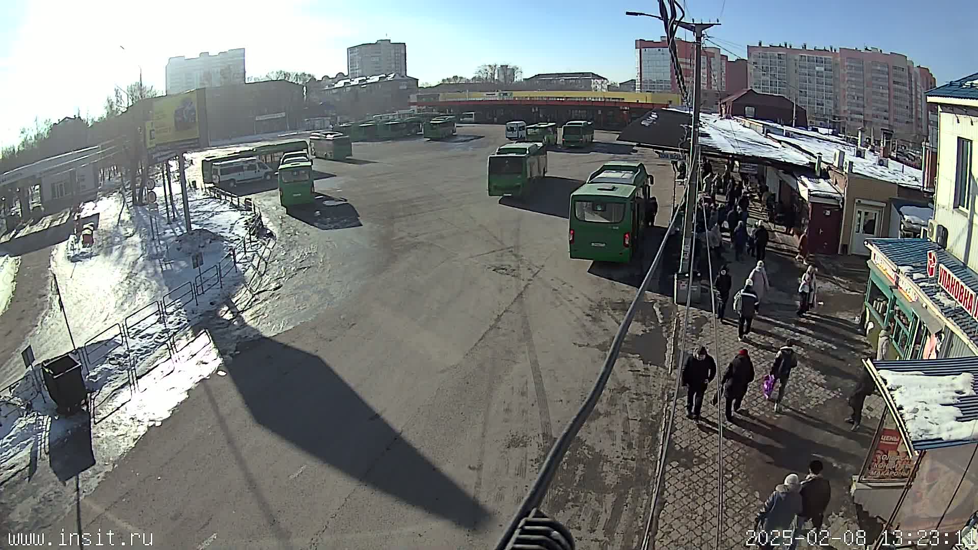 A sunny day at a bus station with numerous green buses parked, and people walking around in a snowy, partially paved area.