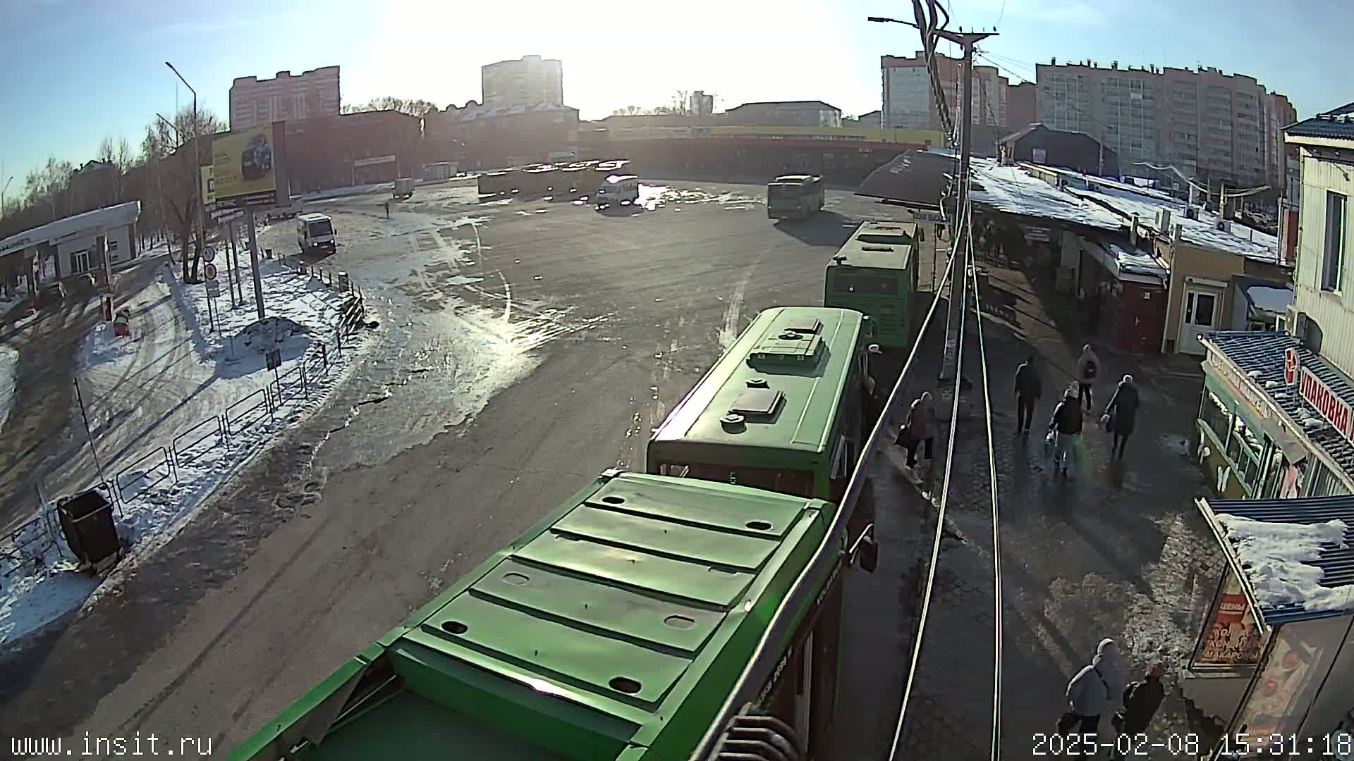 A sunny day reveals a bus station with several green buses, some people walking, and patches of snow and ice on the ground.