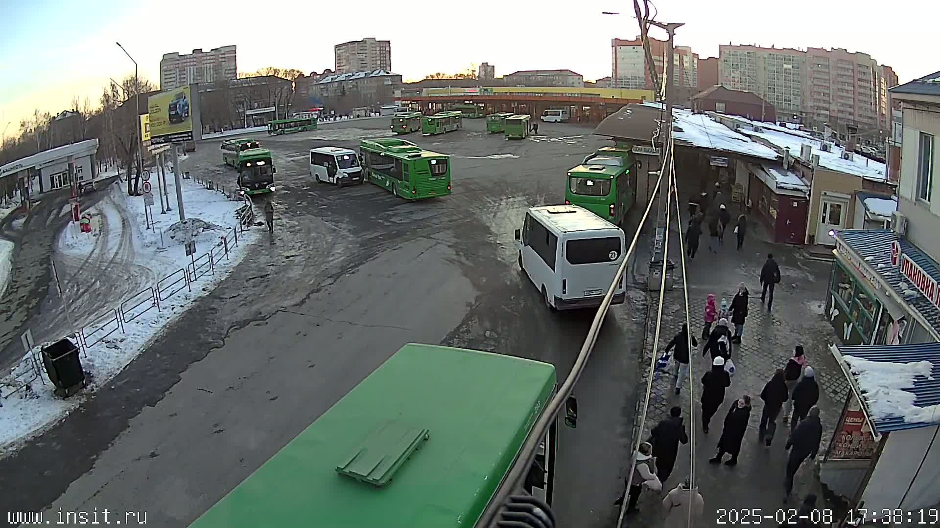 A bus station with several buses and numerous pedestrians, partially covered in snow, under a mostly clear sky at dusk.