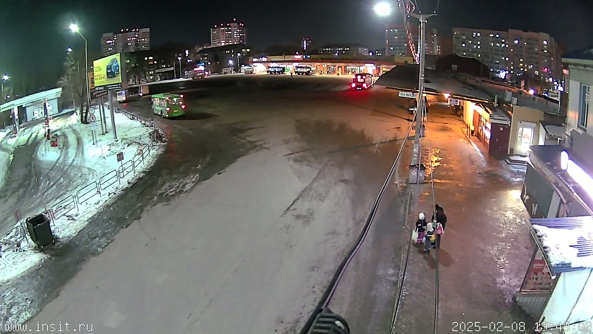A nighttime, snow-covered bus station with several buses parked, a few people walking, and buildings in the background.