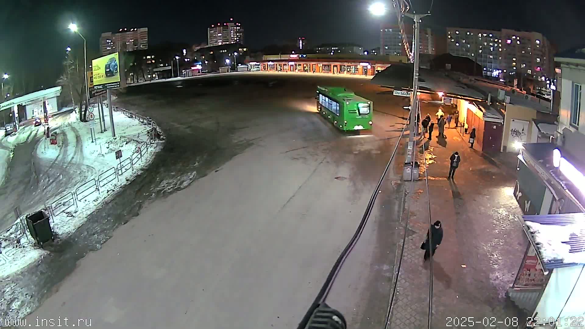 A nighttime, snowy scene shows a green bus at a bus stop with several pedestrians nearby and apartment buildings in the background.