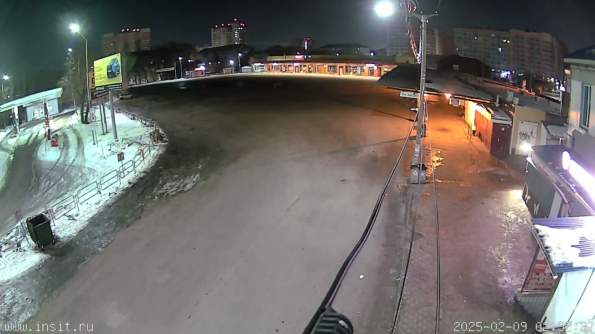 A mostly empty, snow-dusted parking lot and roadway are seen at night, with buildings and streetlights in the background.