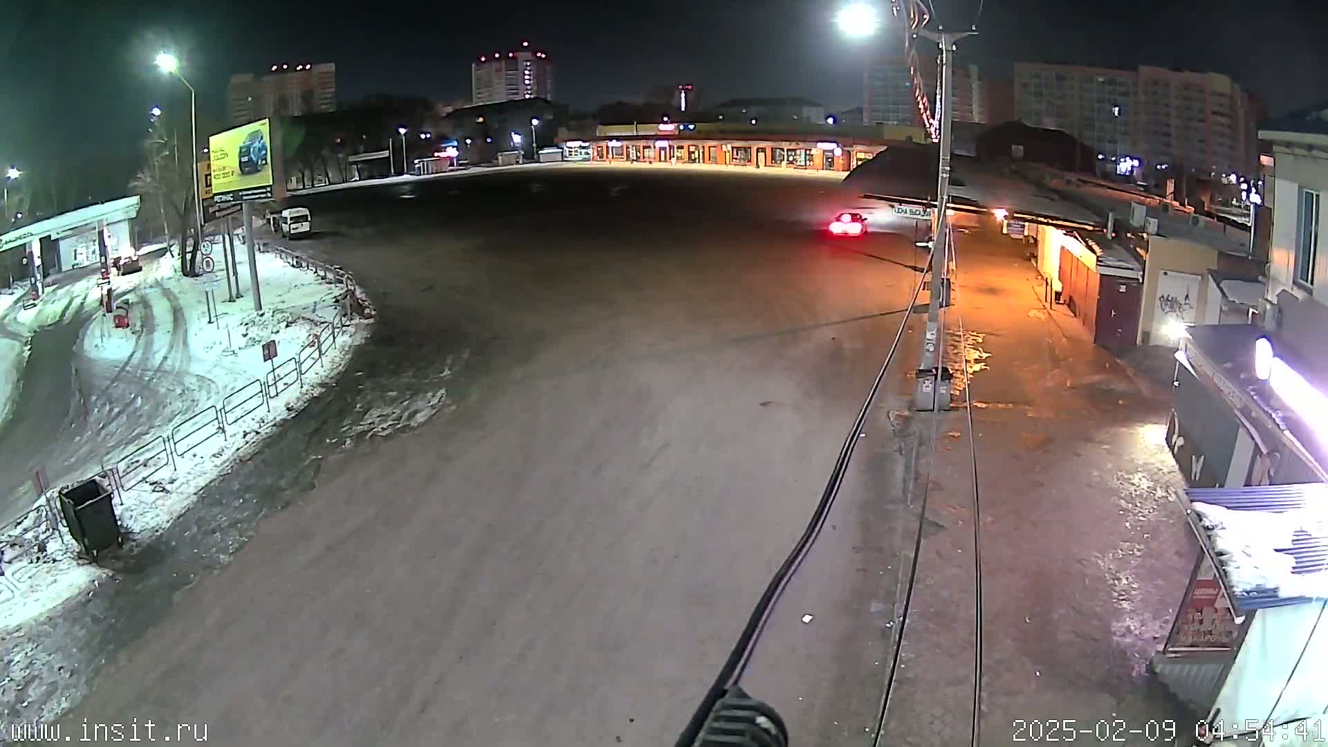 A nighttime, snow-dusted parking lot and roadway are seen, with several cars and buildings in the background under artificial lighting.