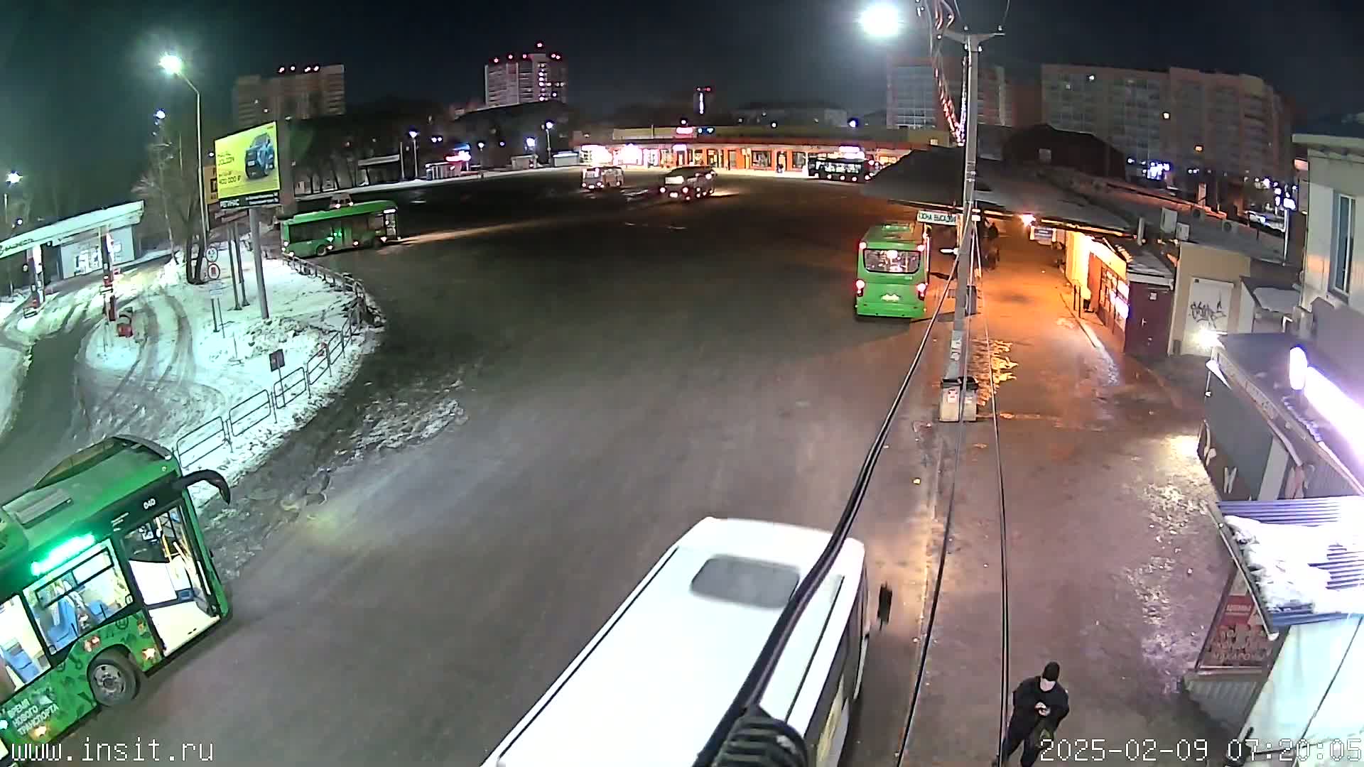 A nighttime view of a bus station with several buses parked and a person walking, amidst light snow on the ground.
