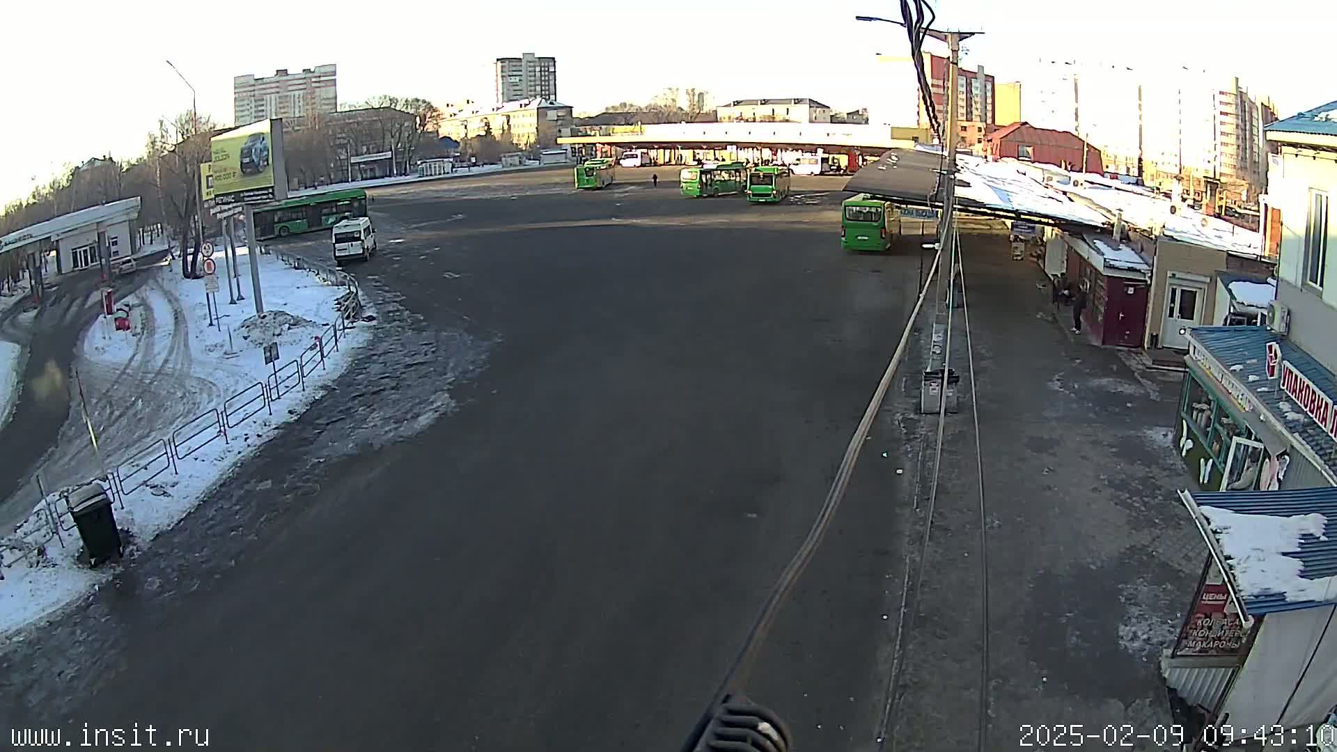 A mostly empty bus station with several green buses parked, surrounded by buildings, under a partly sunny sky with some snow on the ground.