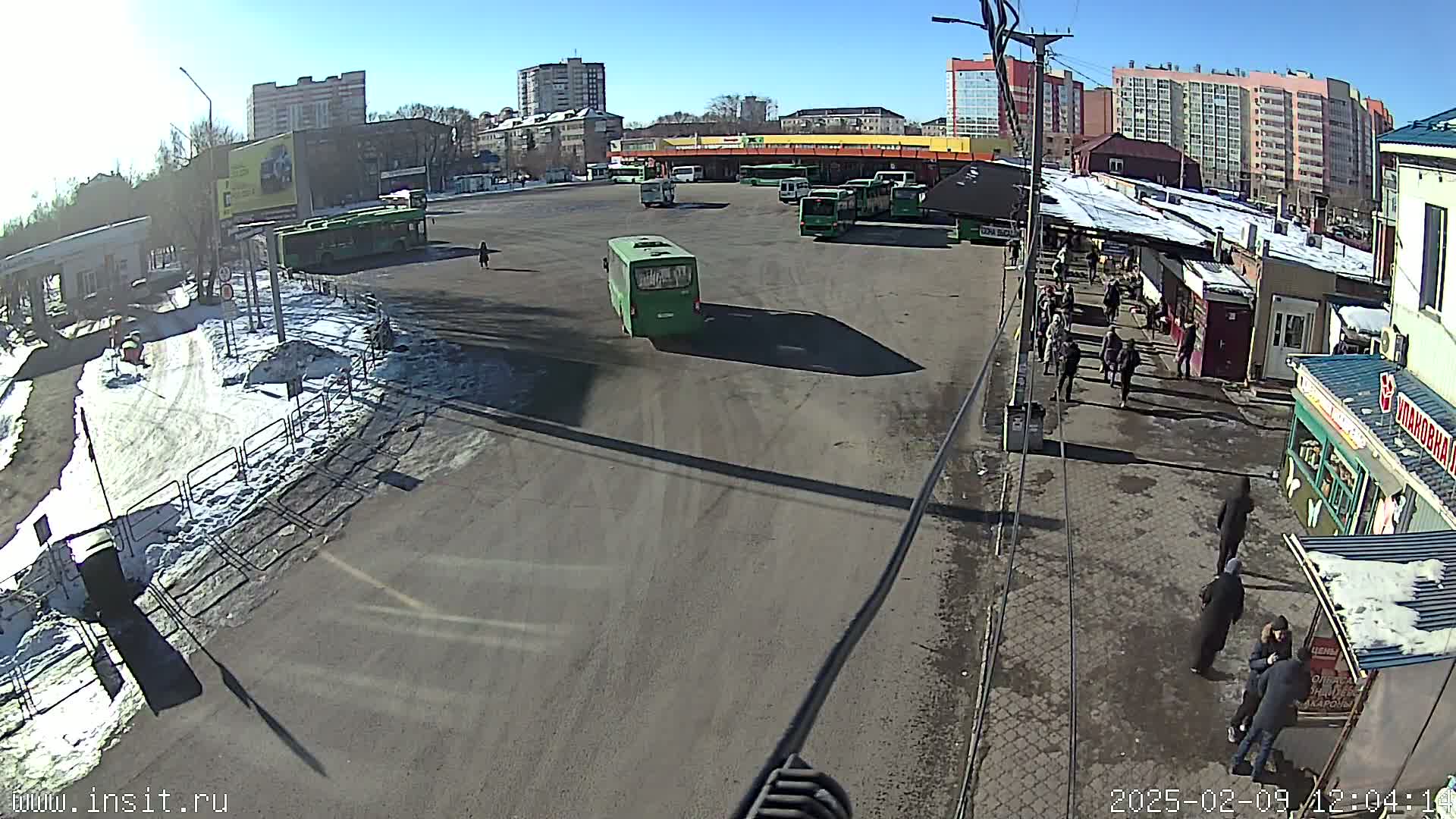 A sunny day at a bus station with several buses parked and people walking around, some snow is visible on the ground.
