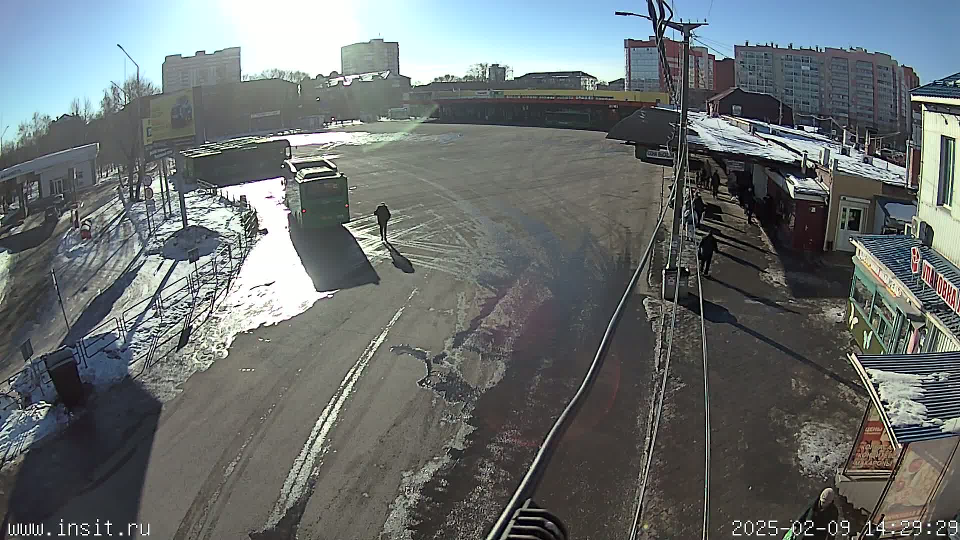 A sunny day reveals a bus station with several buses and people walking around, patches of snow and ice on the ground.