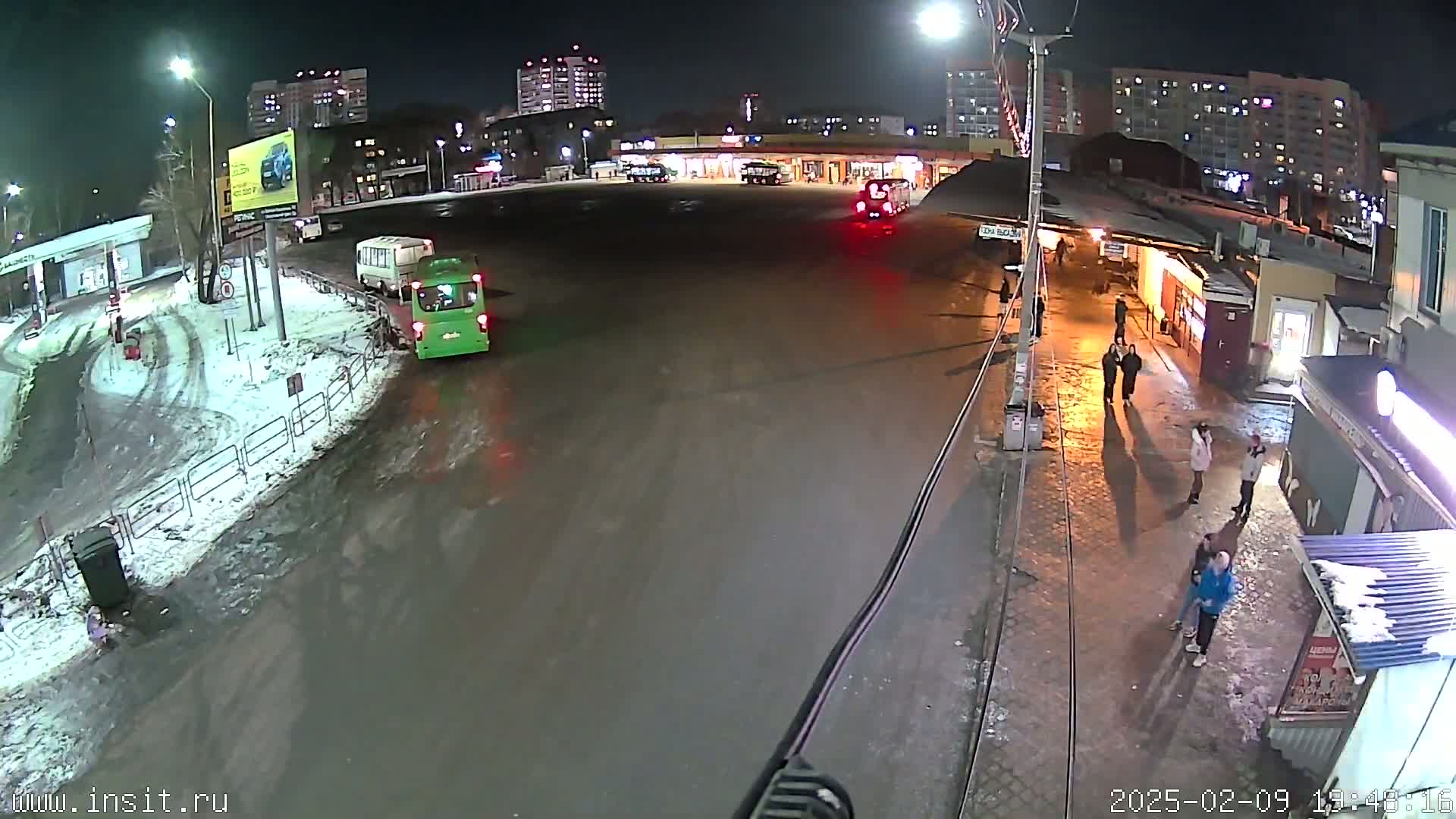 A nighttime view of a bus station with several buses and people present, snow and slush on the ground.