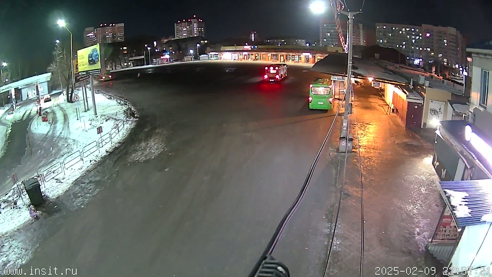 A nighttime view of a bus stop in a snowy area with two buses, one green and one red, and several apartment buildings in the background.