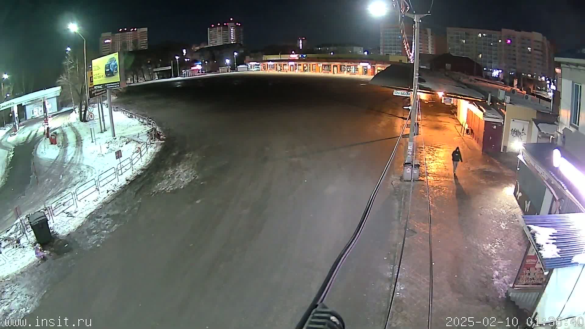 A mostly empty, snow-covered parking lot and roadway at night is illuminated by streetlights, with a single pedestrian walking in the distance.
