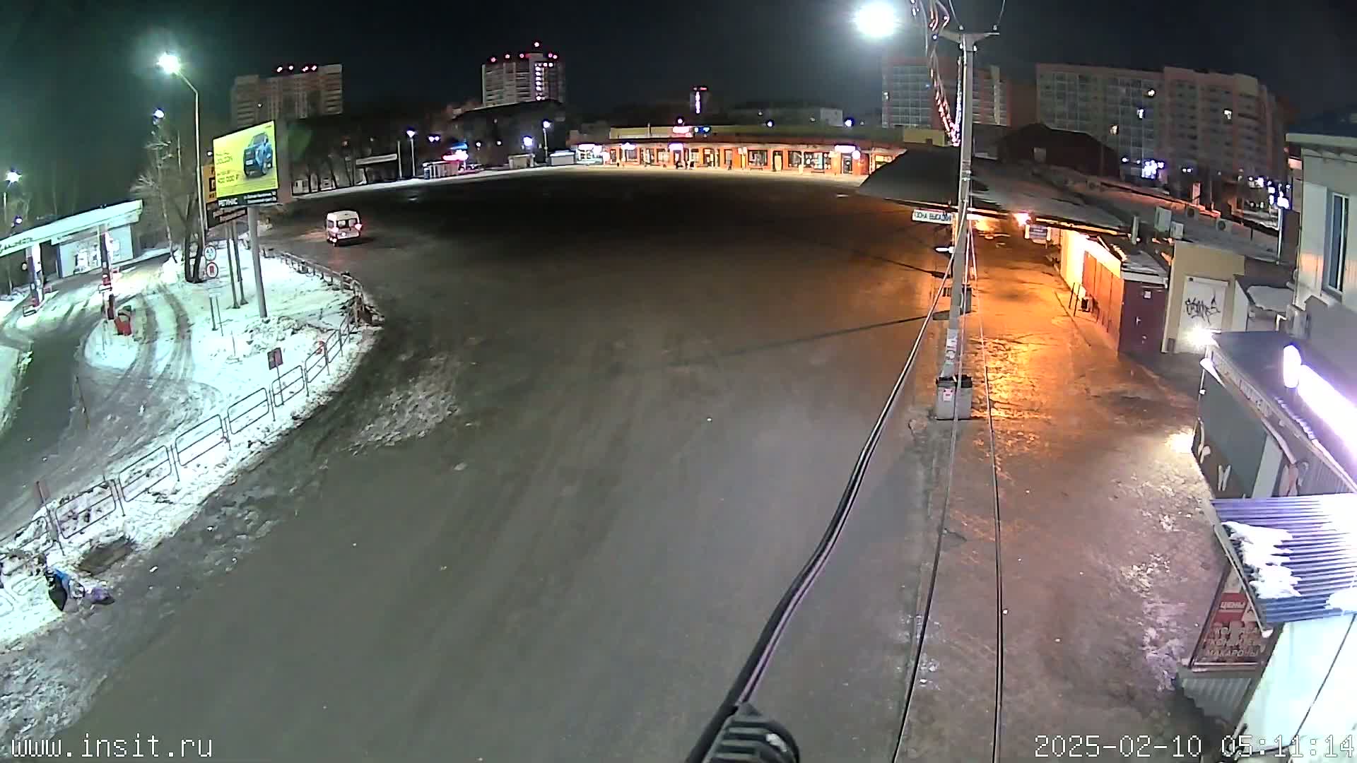 A nighttime, snow-covered parking lot and roadway scene features a few buildings and a single vehicle.