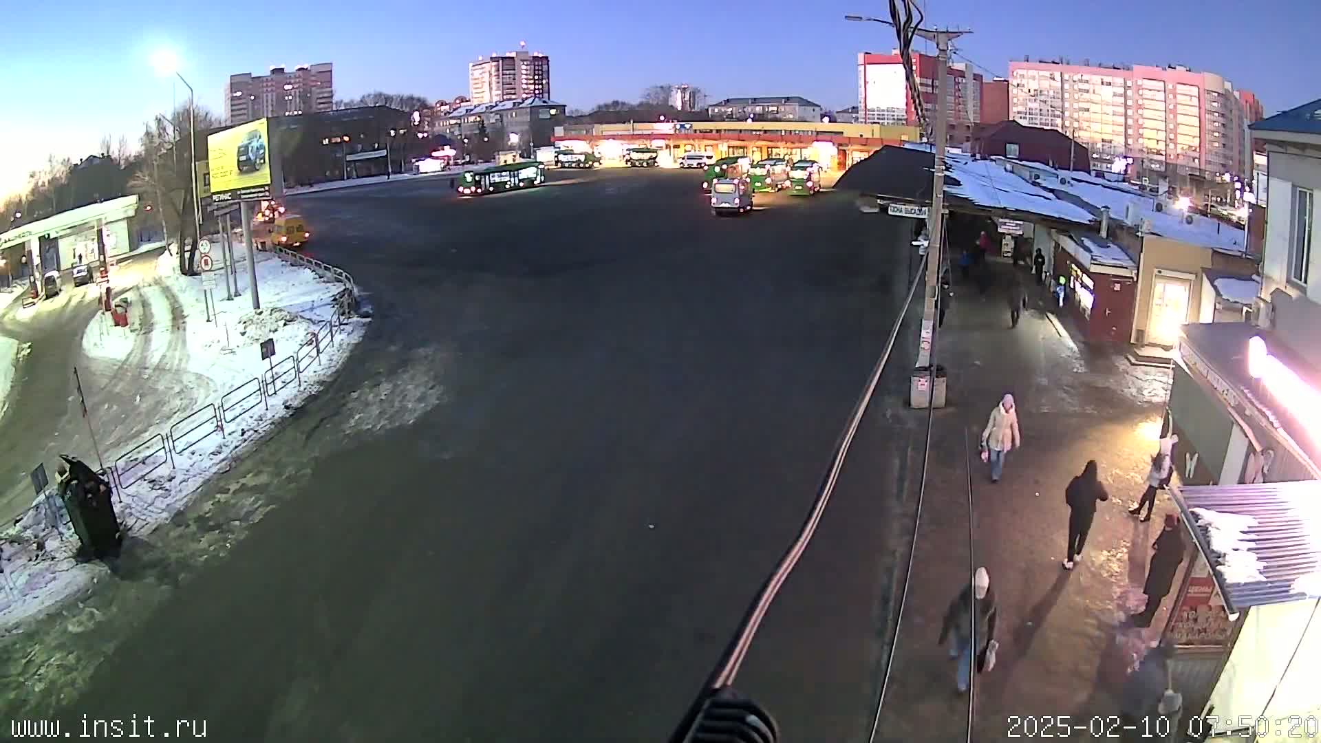 A bus station with numerous buses parked, a few pedestrians walking, and snow on the ground under a twilight sky.
