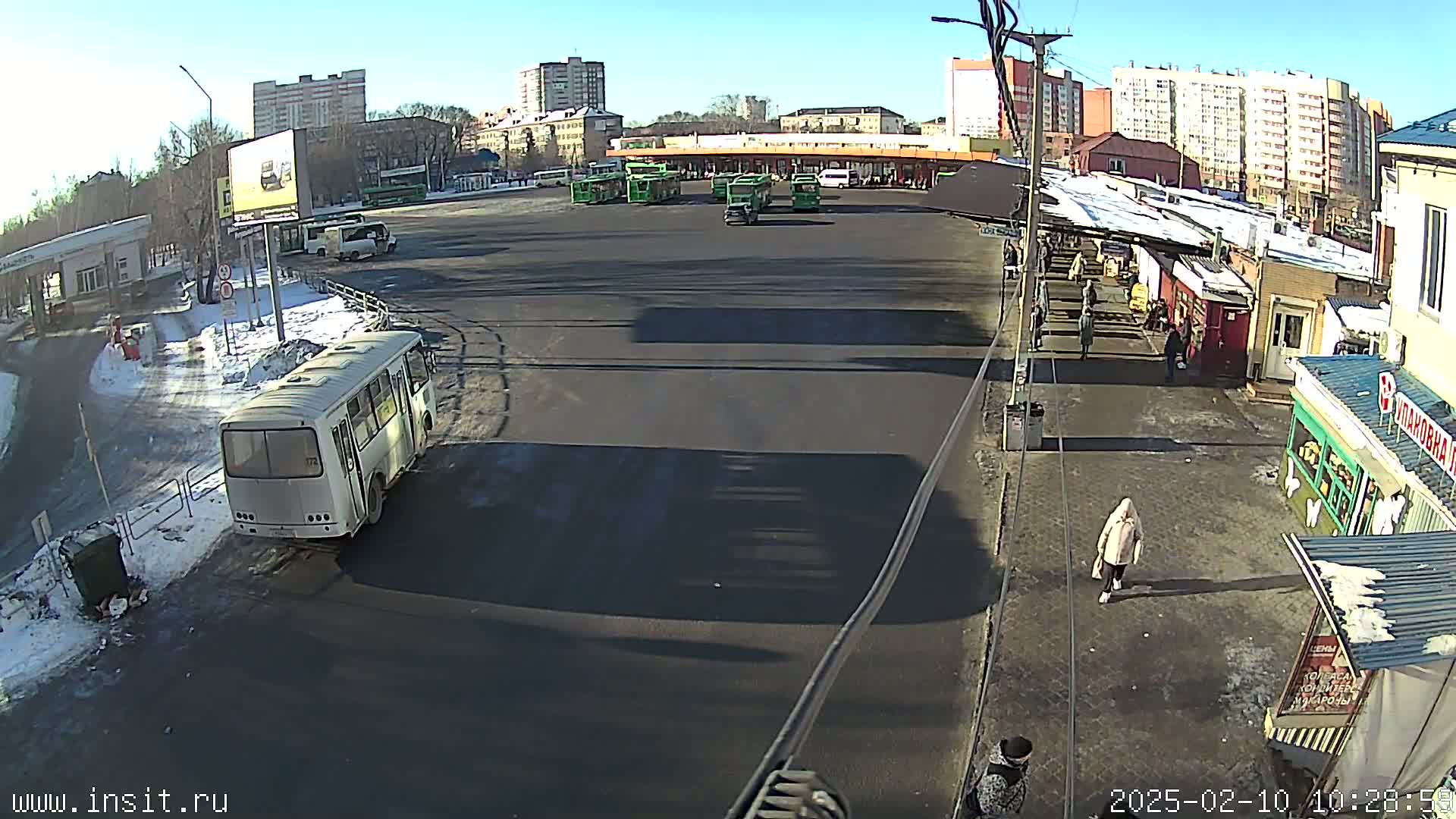 A bus station with several buses parked, surrounded by buildings on a sunny day with patches of snow on the ground.
