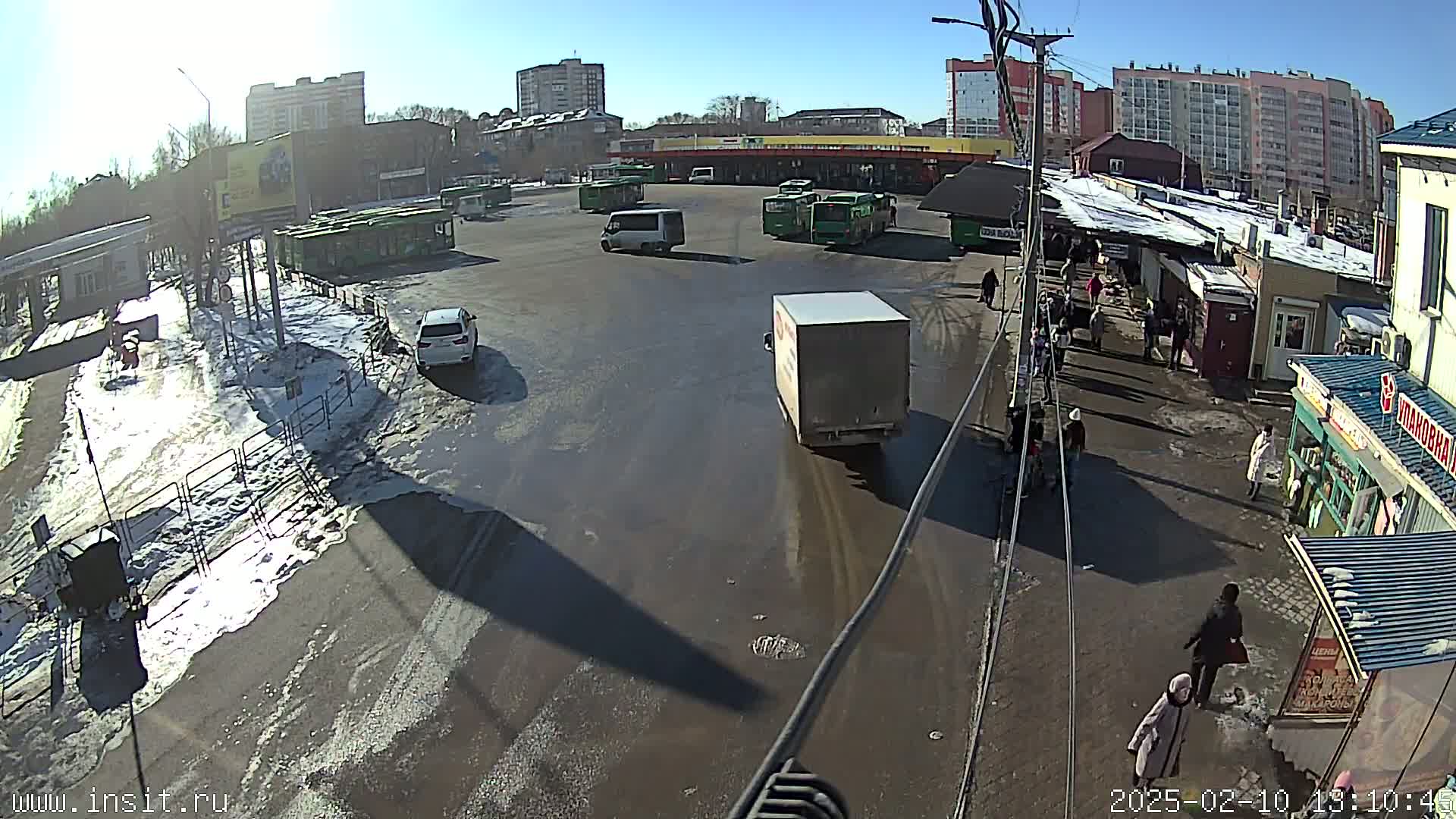 A sunny day reveals a bus station with melting snow and slush on the ground, several buses and vehicles are parked or driving, and people are walking around.