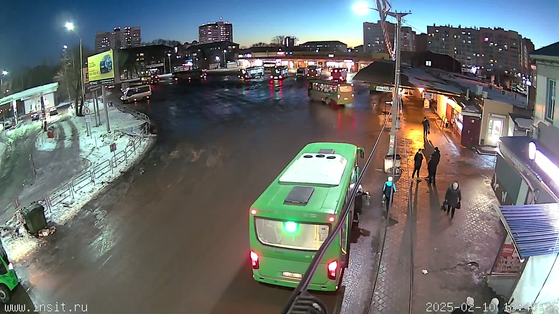 A flooded bus stop at night, with several buses and people present, surrounded by snow and apartment buildings under a dark sky.