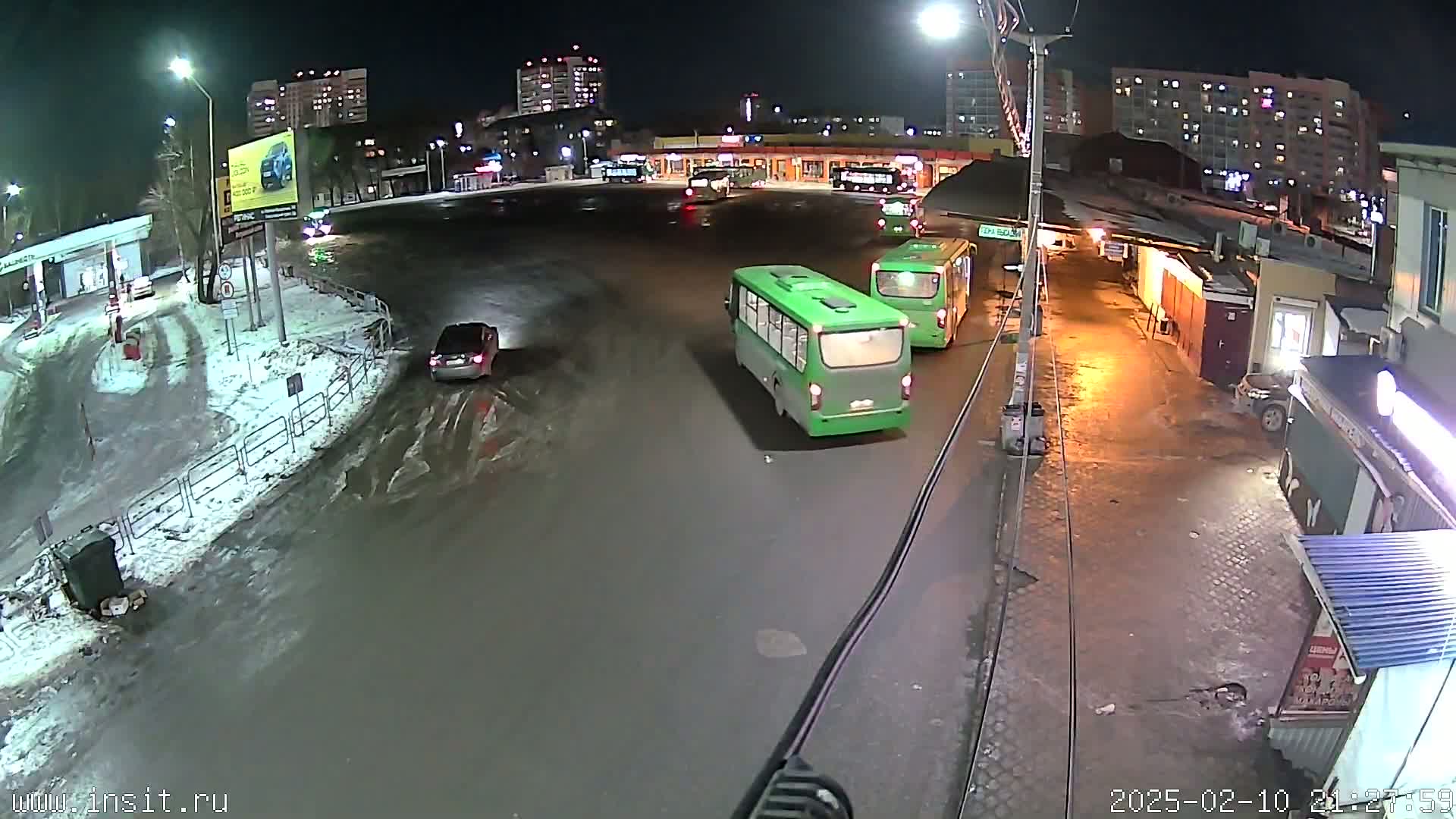 At night, in snowy conditions, a bus station is seen with several buses and cars present.