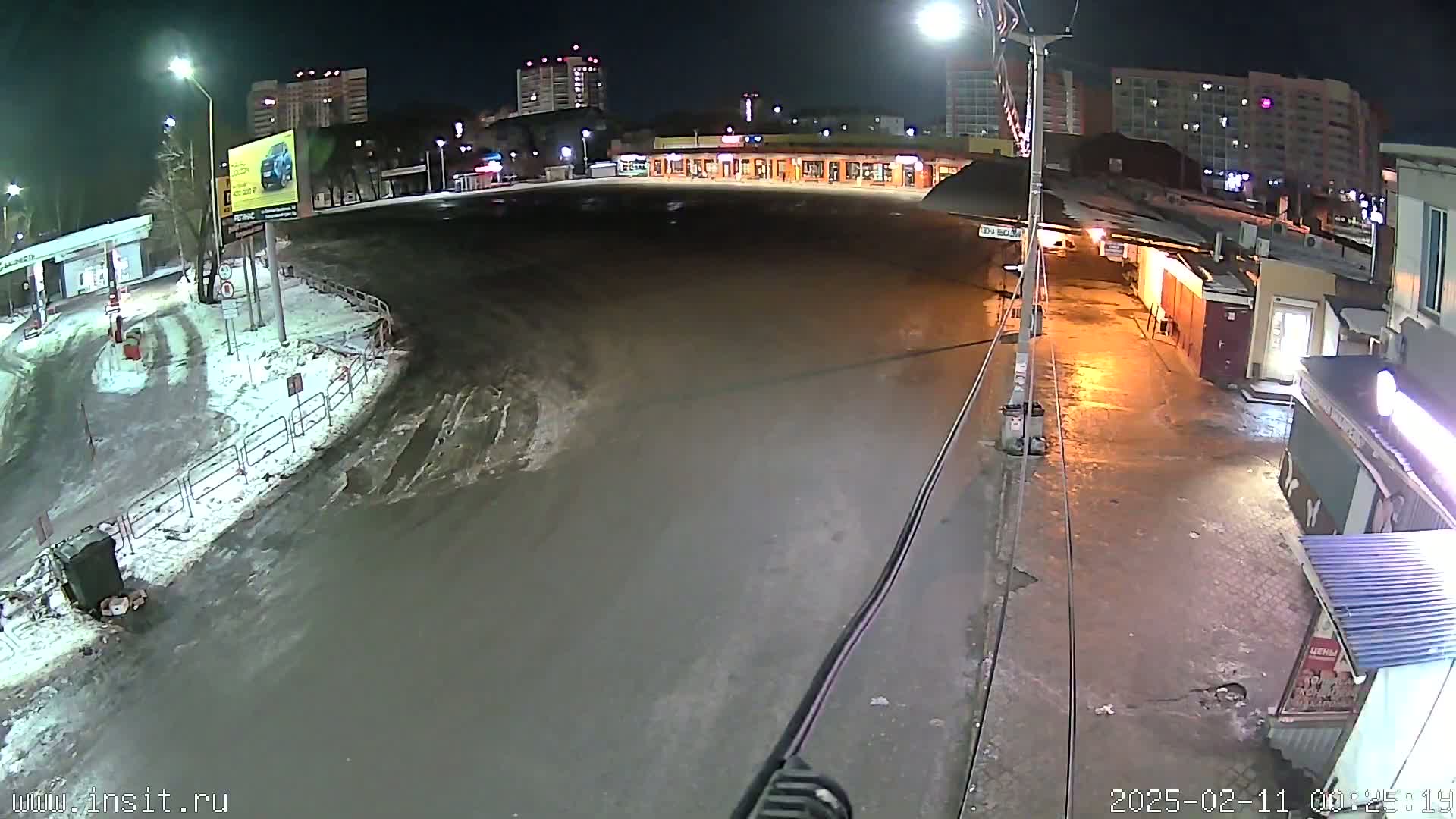 A nighttime, wet, snow-covered street scene shows a mostly empty plaza with surrounding buildings and a gas station.