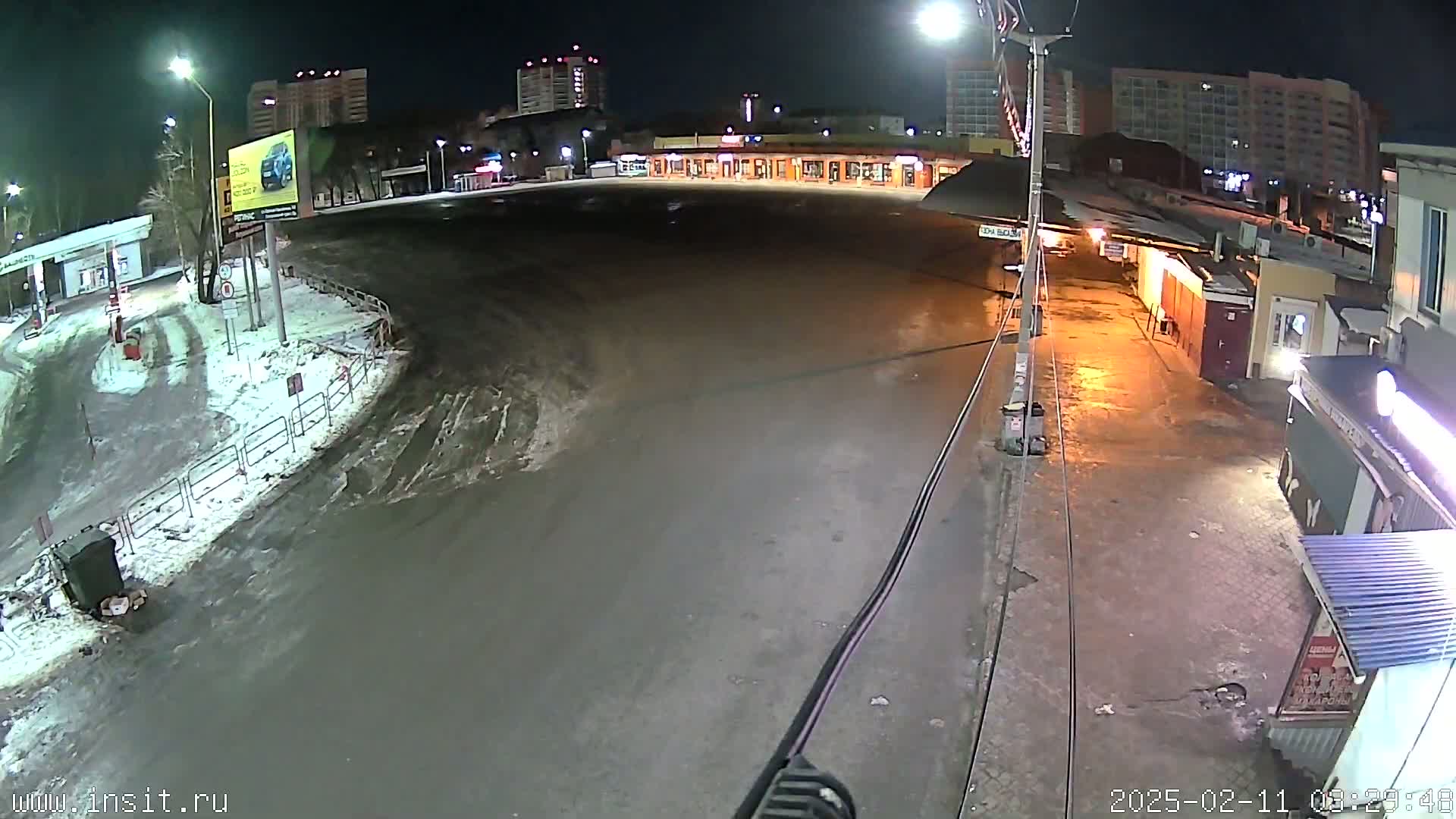 A wet, empty roadway and parking lot at night are illuminated by streetlights, with snow visible along the edges and apartment buildings in the background.