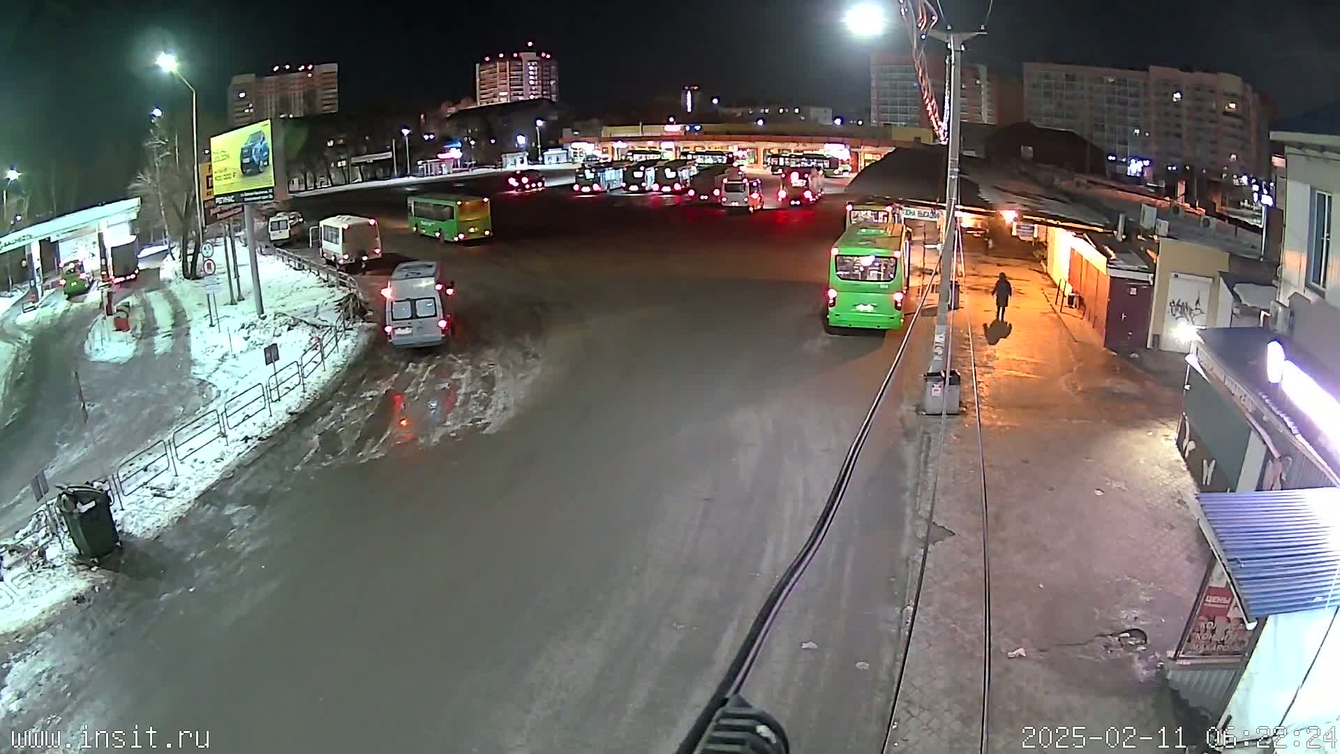 A nighttime, snowy scene shows a bus station with numerous buses parked and a few vehicles on a wet, snow-covered road.