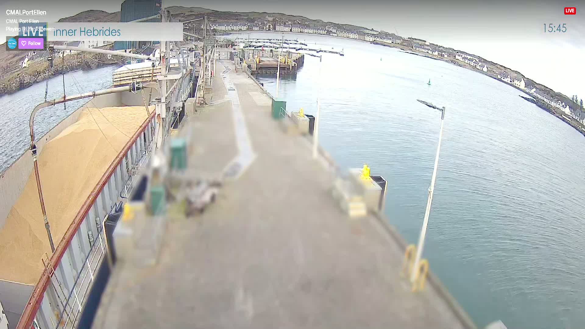 A large cargo ship is being loaded with a tan granular substance at a harbor on a calm, sunny day.