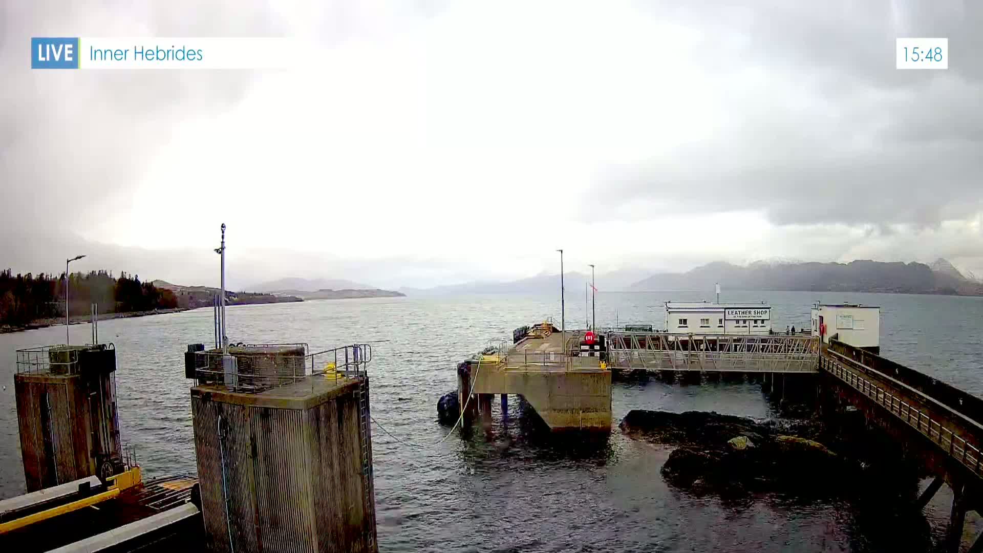 A ferry dock with a small building is seen on a calm, overcast day with mountains in the background.