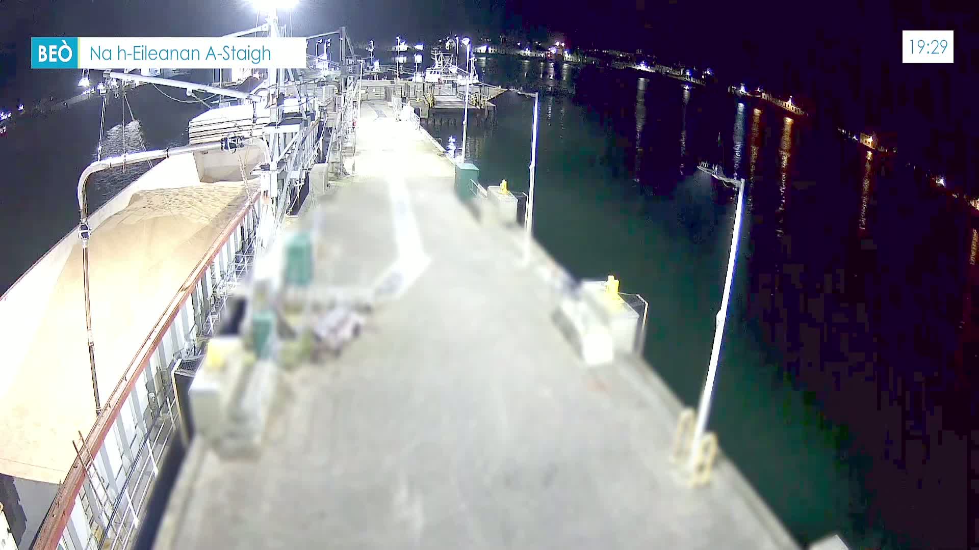 A nighttime view of a dock where a ship is being loaded with a light-brown material under bright lights, with calm dark water visible beyond the dock.